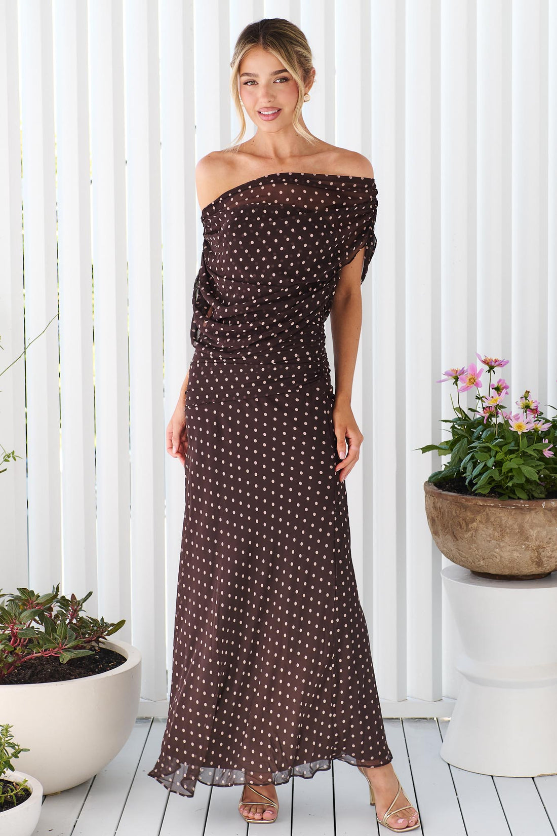 A woman wearing the minnie + MAXXIE Pinto off-shoulder, maxi dark brown dress with polka dots, standing in front of a white vertical panelled wall.