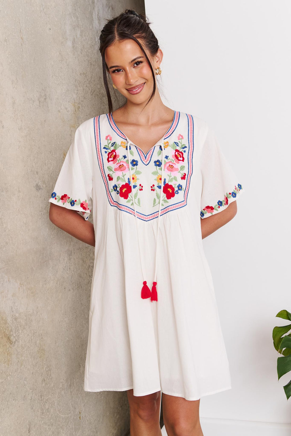 A woman wearing the minnie + MAXXIE Posie cotton dress, a white short-sleeve tunic dress with colourful floral embroidery around the neckline and sleeve cuffs, standing against a concrete and white wall.