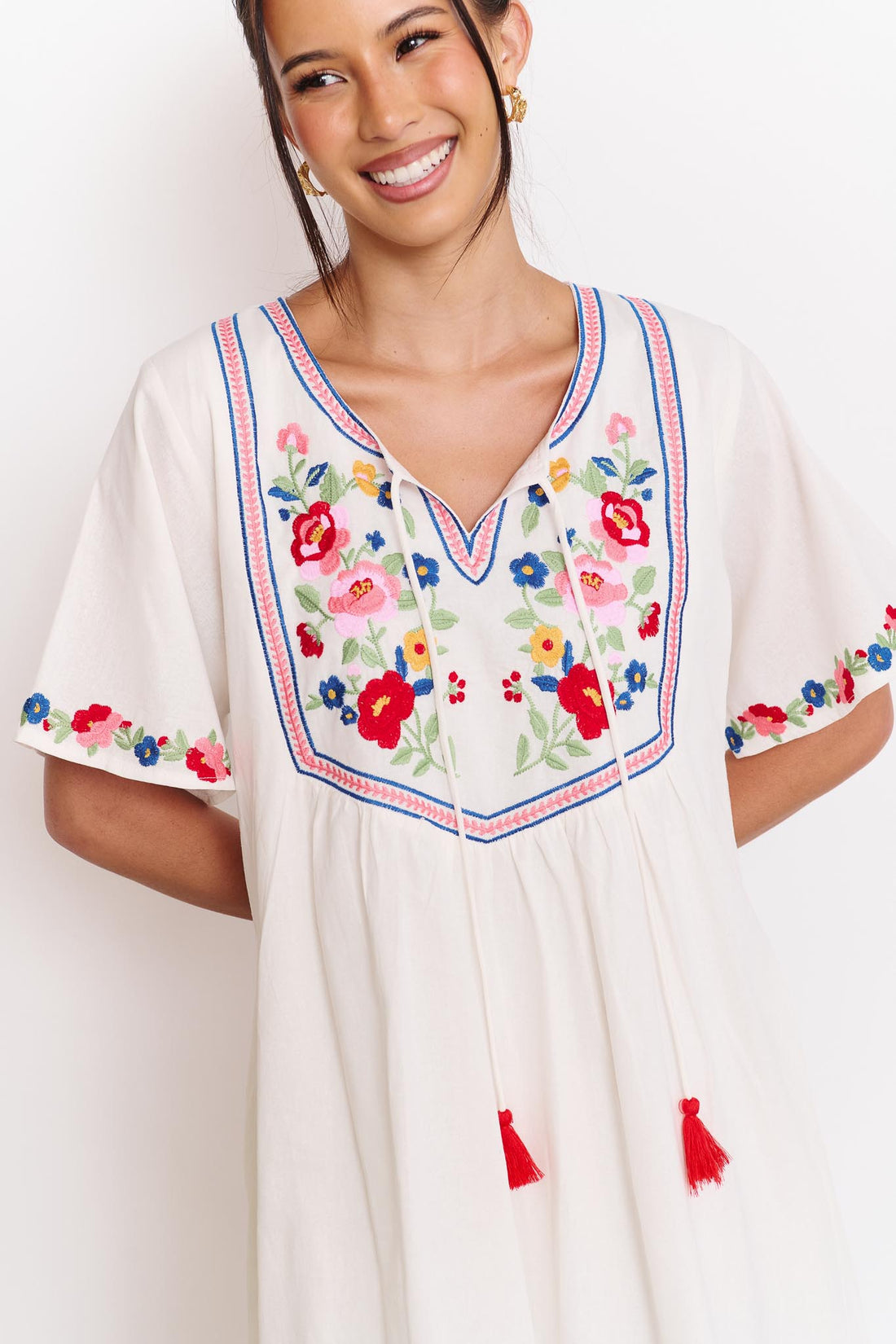 Detail of a woman wearing the minnie + MAXXIE Posie cotton dress, a white short-sleeve tunic dress with colourful floral embroidery around the neckline and sleeve cuffs, and tassle ties at the neckline.