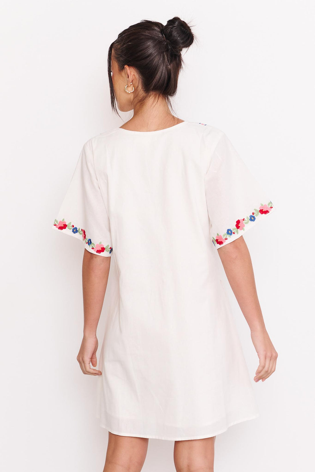 Back view of Aa woman wearing the minnie + MAXXIE Posie cotton dress, a white short-sleeve tunic dress with colourful floral embroidery around the sleeve cuffs, standing against a white wall.