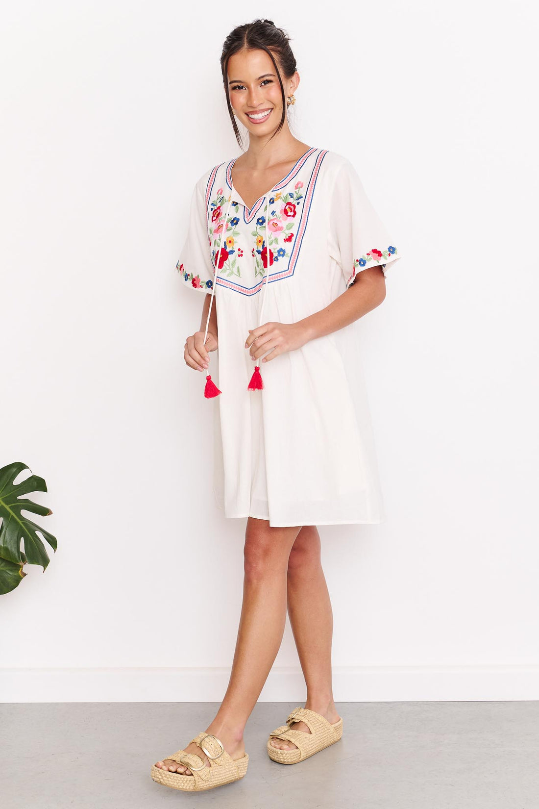 A woman wearing the minnie + MAXXIE Posie cotton dress, a white short-sleeve tunic dress with colourful floral embroidery around the neckline and sleeve cuffs, paired with tan slide sandals, standing against a white wall.