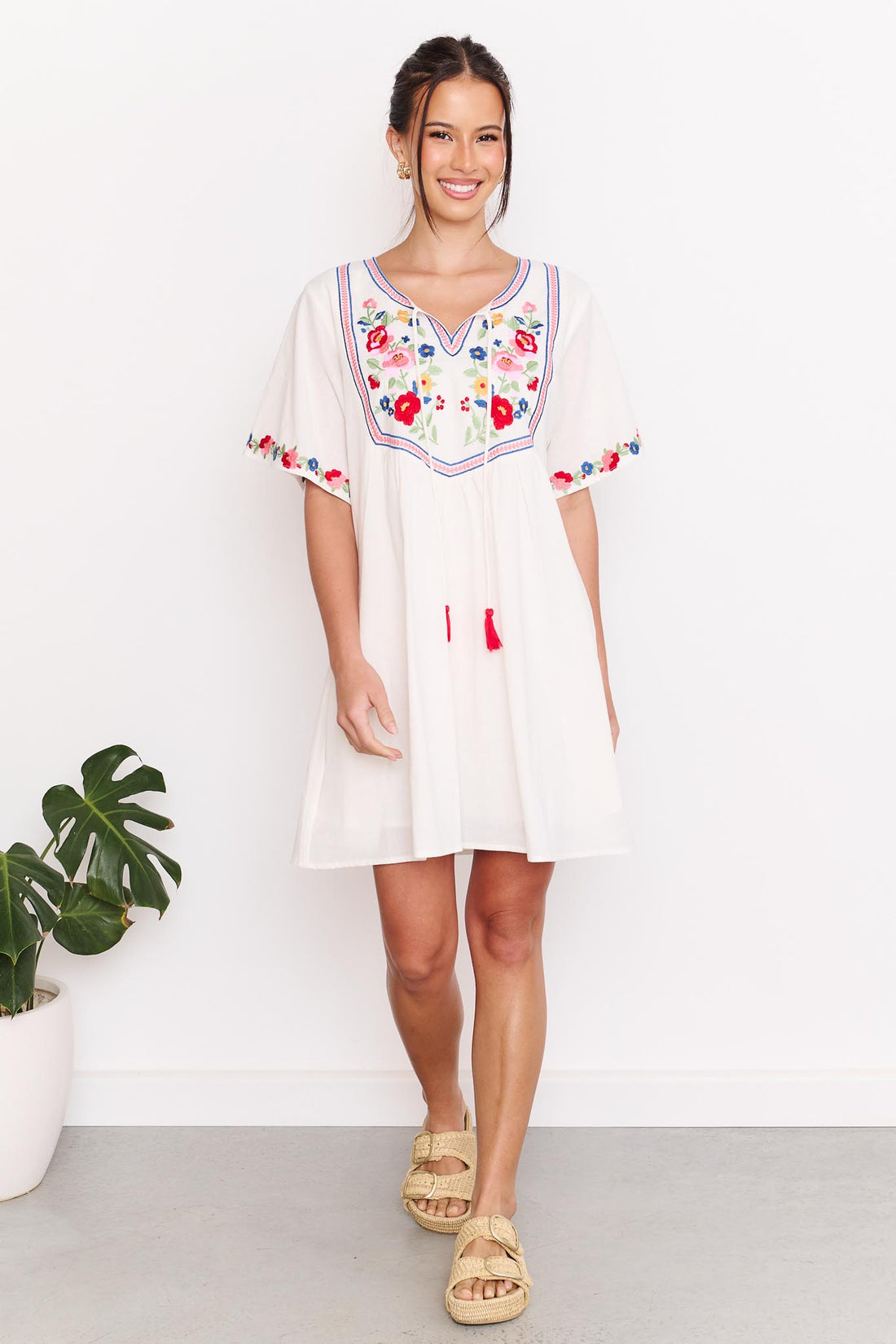 A woman wearing the minnie + MAXXIE Posie cotton dress, a white short-sleeve tunic dress with colourful floral embroidery around the neckline and sleeve cuffs, paired with tan slide sandals, standing against a white wall.
