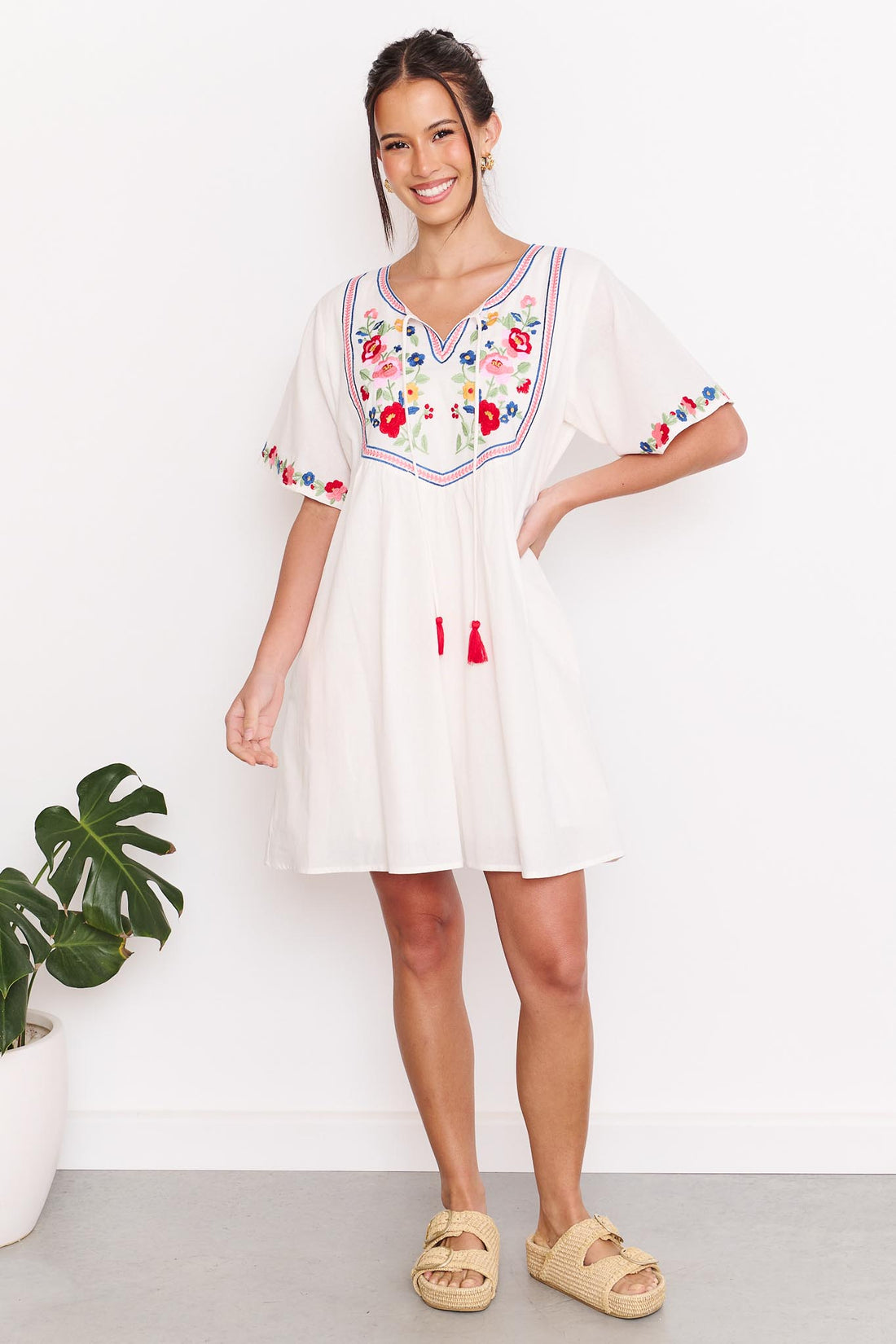 A woman wearing the minnie + MAXXIE Posie cotton dress, a white short-sleeve tunic dress with colourful floral embroidery around the neckline and sleeve cuffs, paired with tan slide sandals, standing against a white wall.