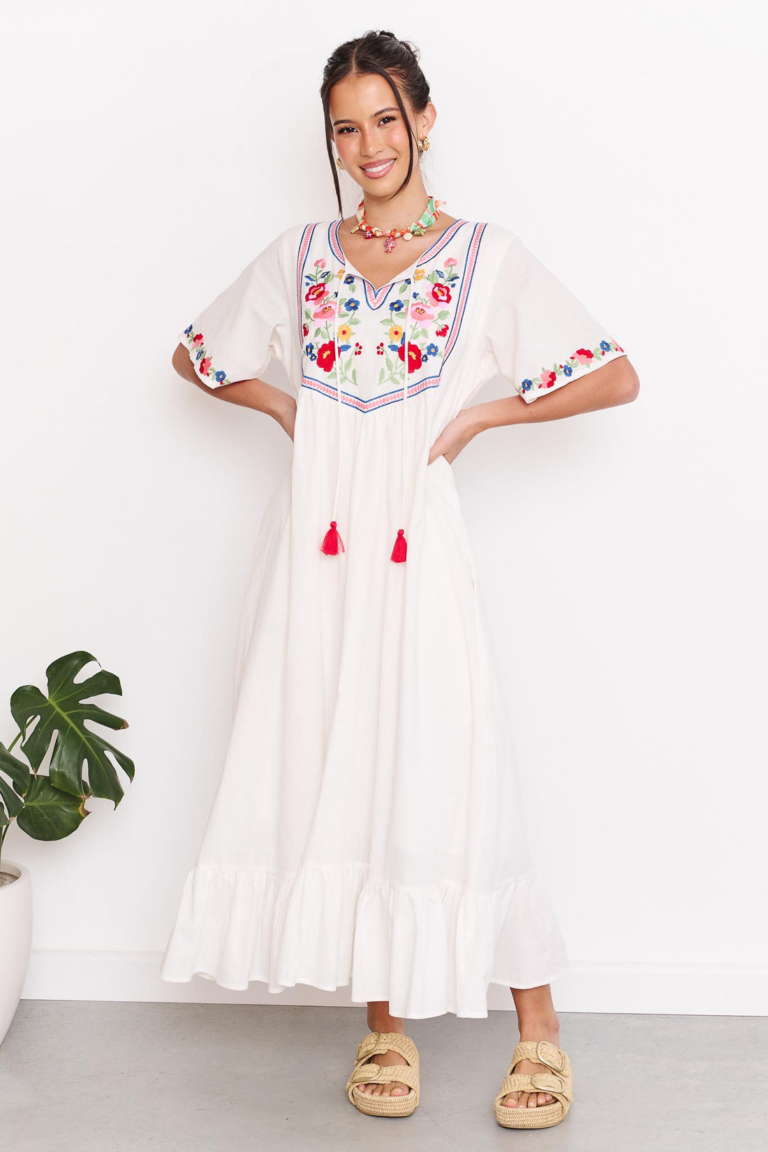 Woman wearing the minnie + MAXXIE Posie Cotton Maxi Dress, a white cotton maxi dress with colourful floral embroidery, V-neckline and short sleeves, styled with the Boheme Charm Necklace and tan slide sandals.