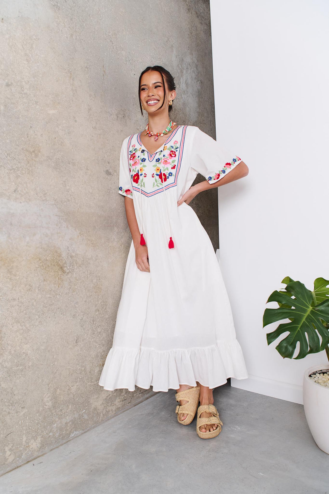 Woman wearing the minnie + MAXXIE Posie Cotton Maxi Dress, a white cotton maxi dress with colourful floral embroidery, V-neckline and short sleeves, styled with the Boheme Charm Necklace and tan slide sandals.