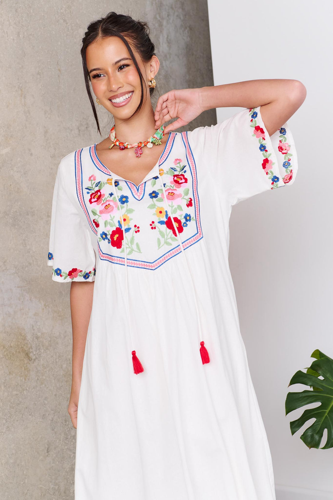 Woman wearing the minnie + MAXXIE Posie Cotton Maxi Dress, a white cotton maxi dress with colourful floral embroidery, V-neckline and short sleeves, styled with the Boheme Charm Necklace.