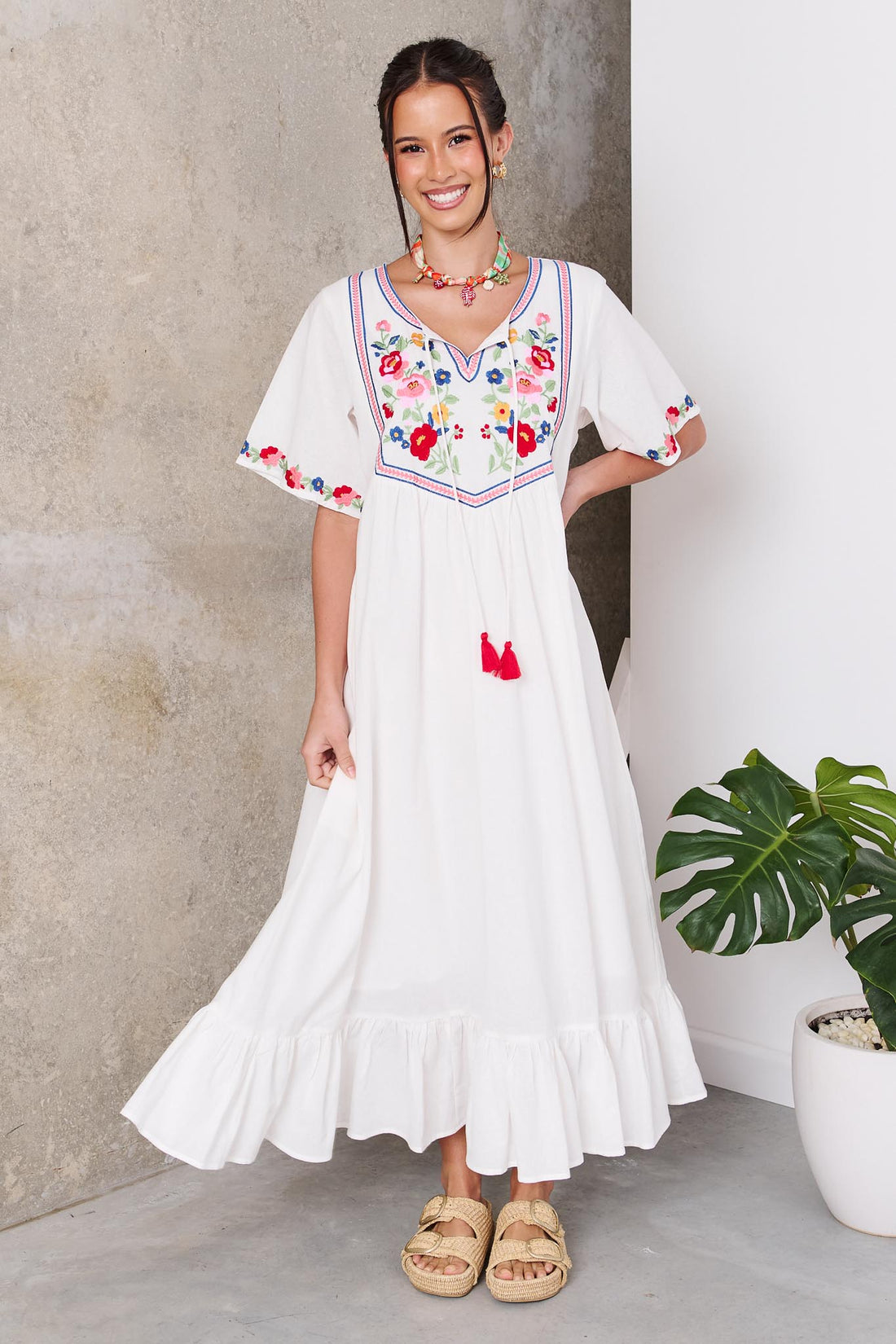 Woman wearing the minnie + MAXXIE Posie Cotton Maxi Dress, a white cotton maxi dress with colourful floral embroidery, V-neckline and short sleeves, styled with the Boheme Charm Necklace and tan slide sandals.