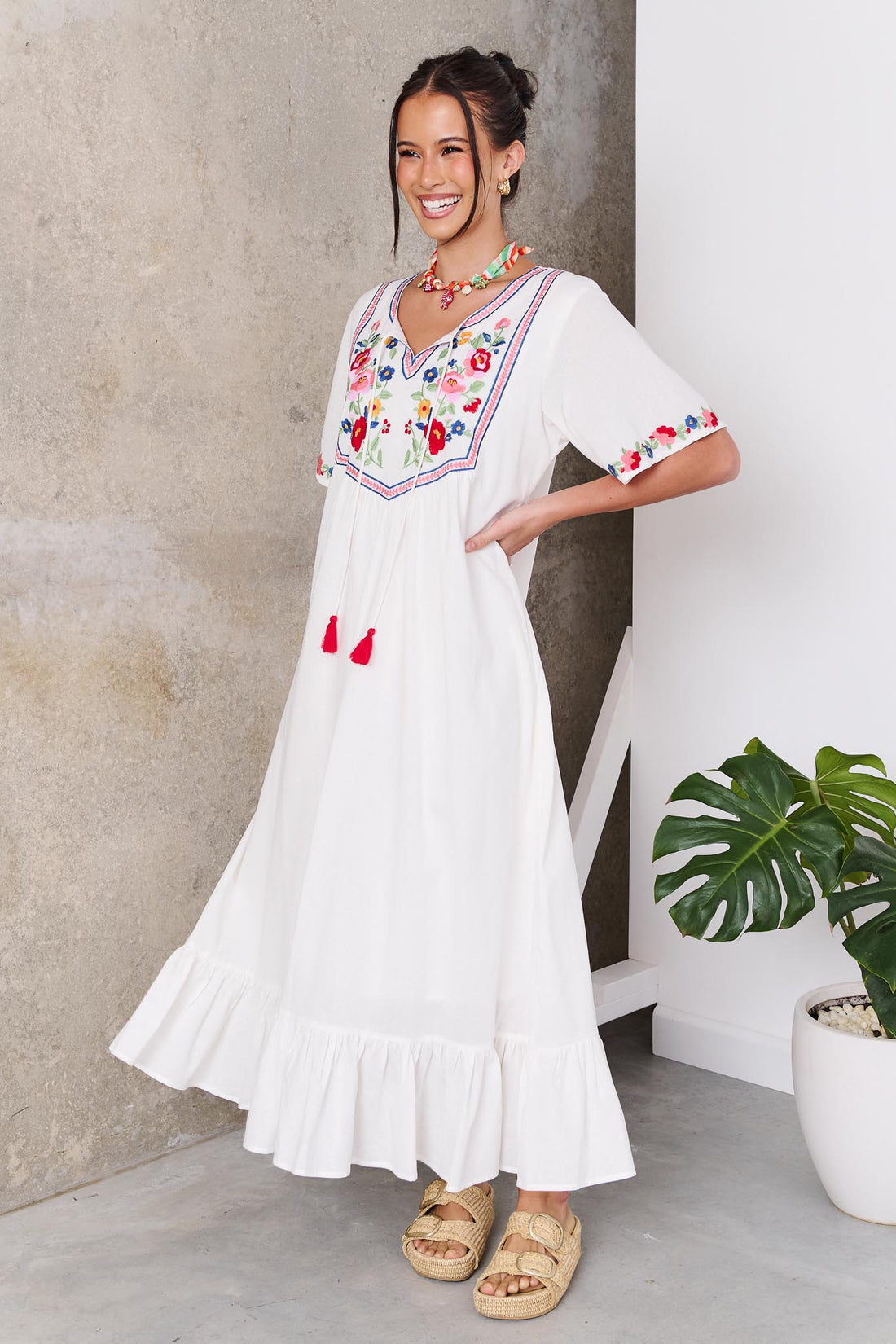Woman wearing the minnie + MAXXIE Posie Cotton Maxi Dress, a white cotton maxi dress with colourful floral embroidery, V-neckline and short sleeves, styled with the Boheme Charm Necklace and tan slide sandals.