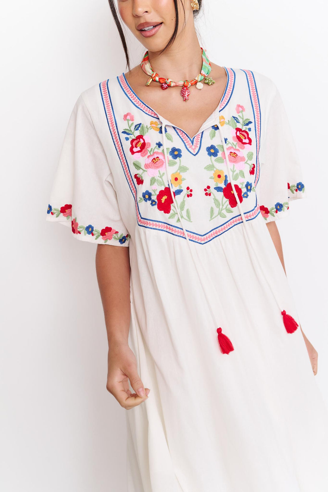 Detail of a woman wearing the minnie + MAXXIE Posie Cotton Maxi Dress, a white cotton maxi dress with colourful floral embroidery, V-neckline and short sleeves, styled with the Boheme Charm Necklace.