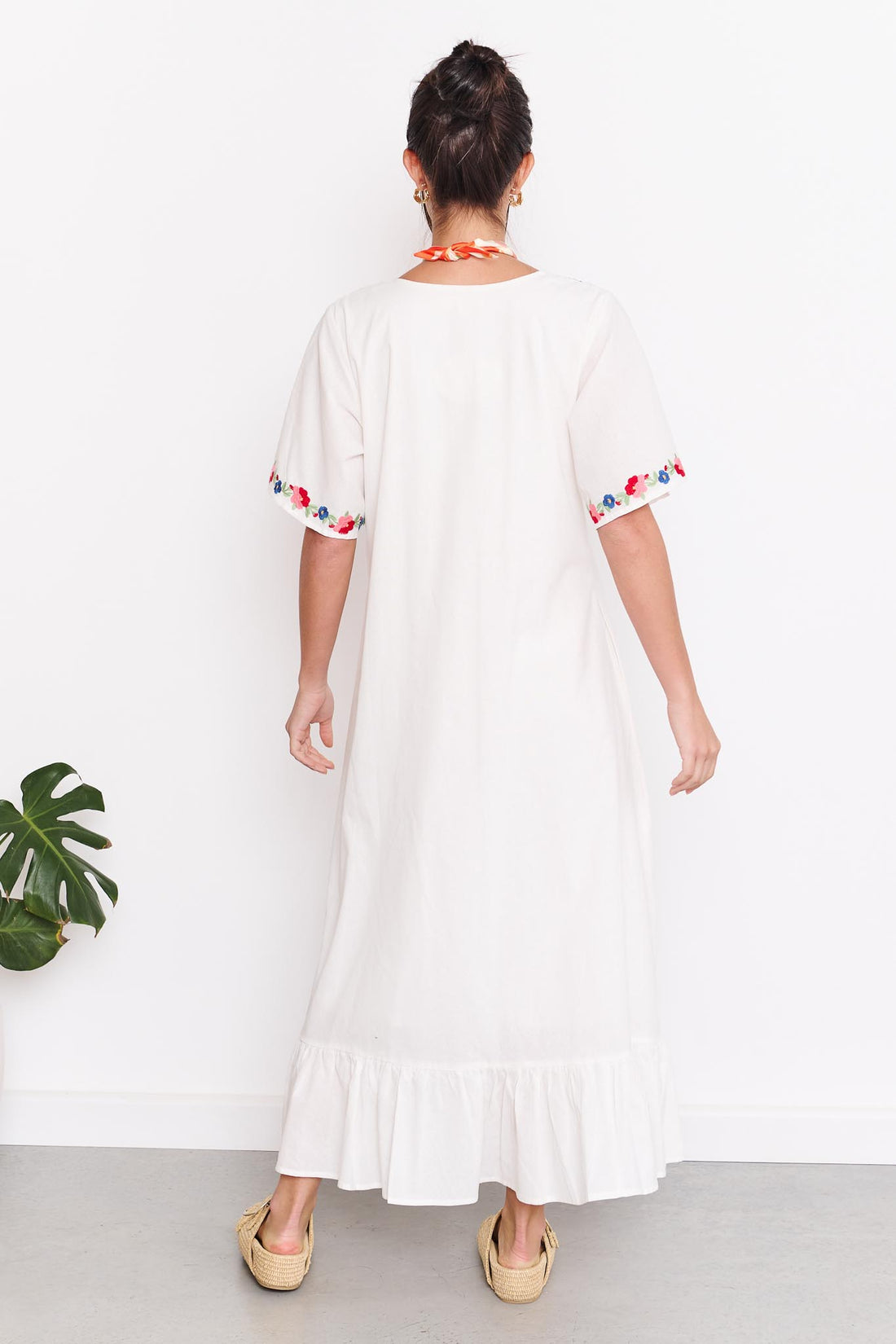 Back view of a woman wearing the minnie + MAXXIE Posie Cotton Maxi Dress, a white cotton maxi dress with colourful floral embroidery, V-neckline and short sleeves, styled with the Boheme Charm Necklace and tan slide sandals.
