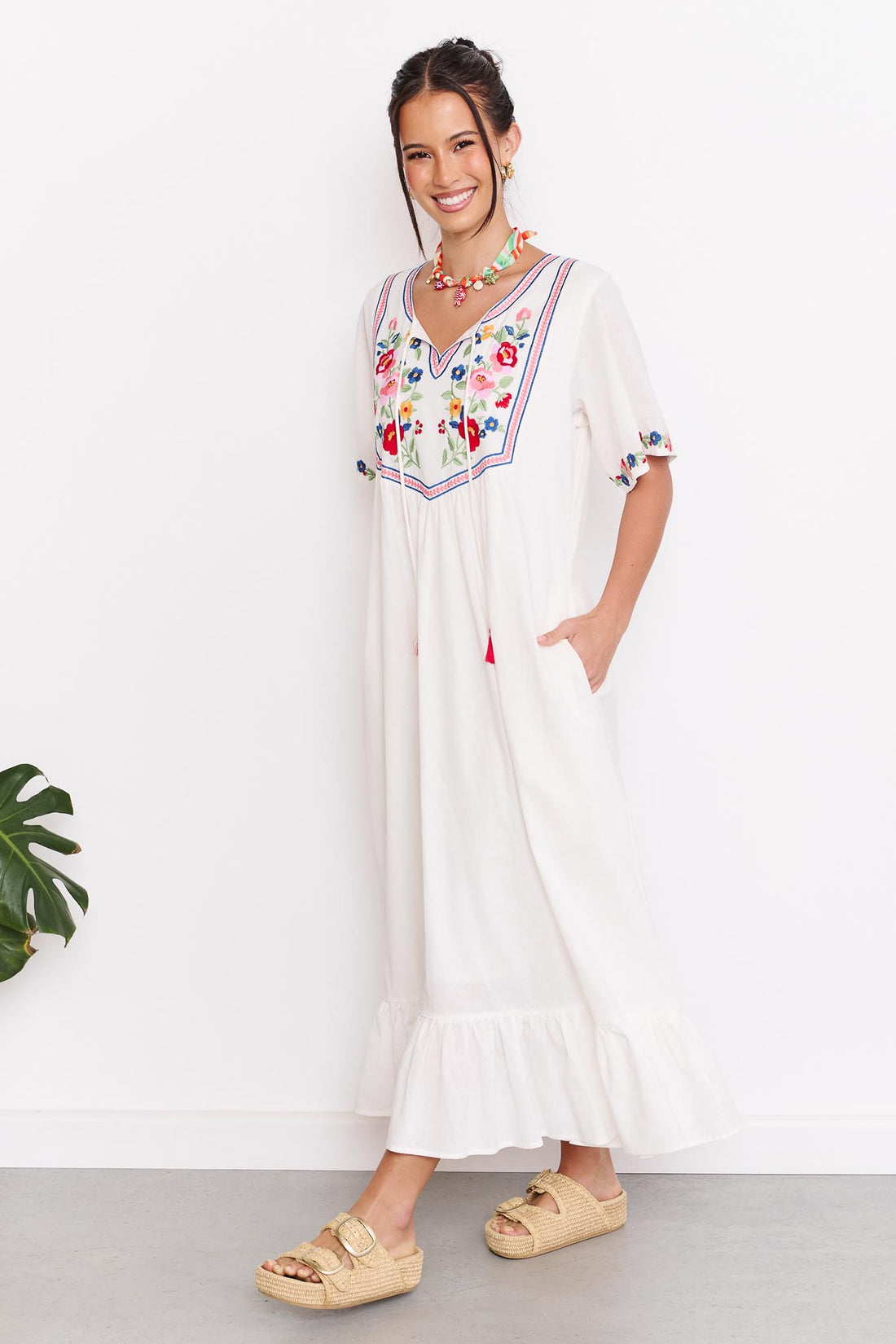 Woman wearing the minnie + MAXXIE Posie Cotton Maxi Dress, a white cotton maxi dress with colourful floral embroidery, V-neckline and short sleeves, styled with the Boheme Charm Necklace and tan slide sandals.