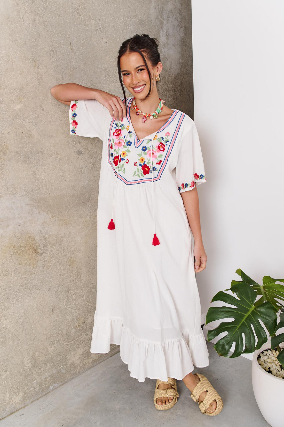 Woman wearing the minnie + MAXXIE Posie Cotton Maxi Dress, a white cotton maxi dress with colourful floral embroidery, V-neckline and short sleeves, styled with the Boheme Charm Necklace and tan slide sandals.