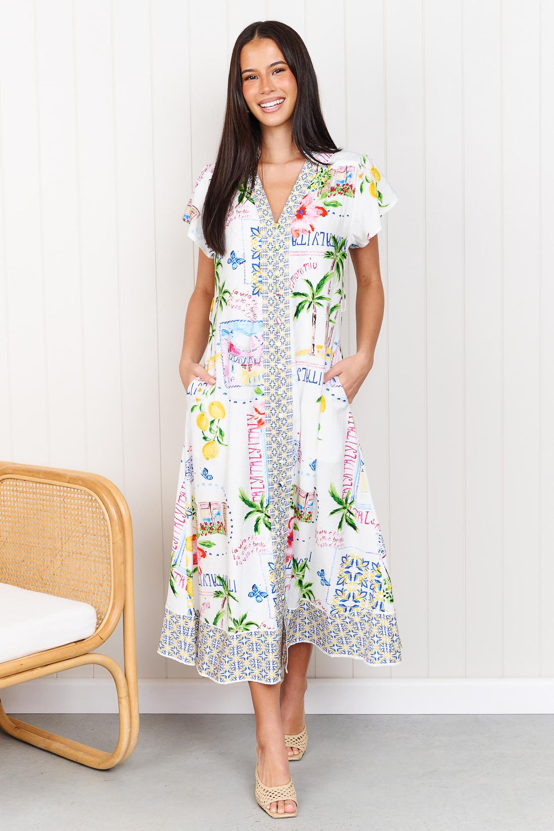 A woman wearing the minnie + MAXXIE Positano Midi Dress, featuring a colourful mediterranean-themed print, short flutter sleeves, and a V-neckline, set against a white panelled wall.