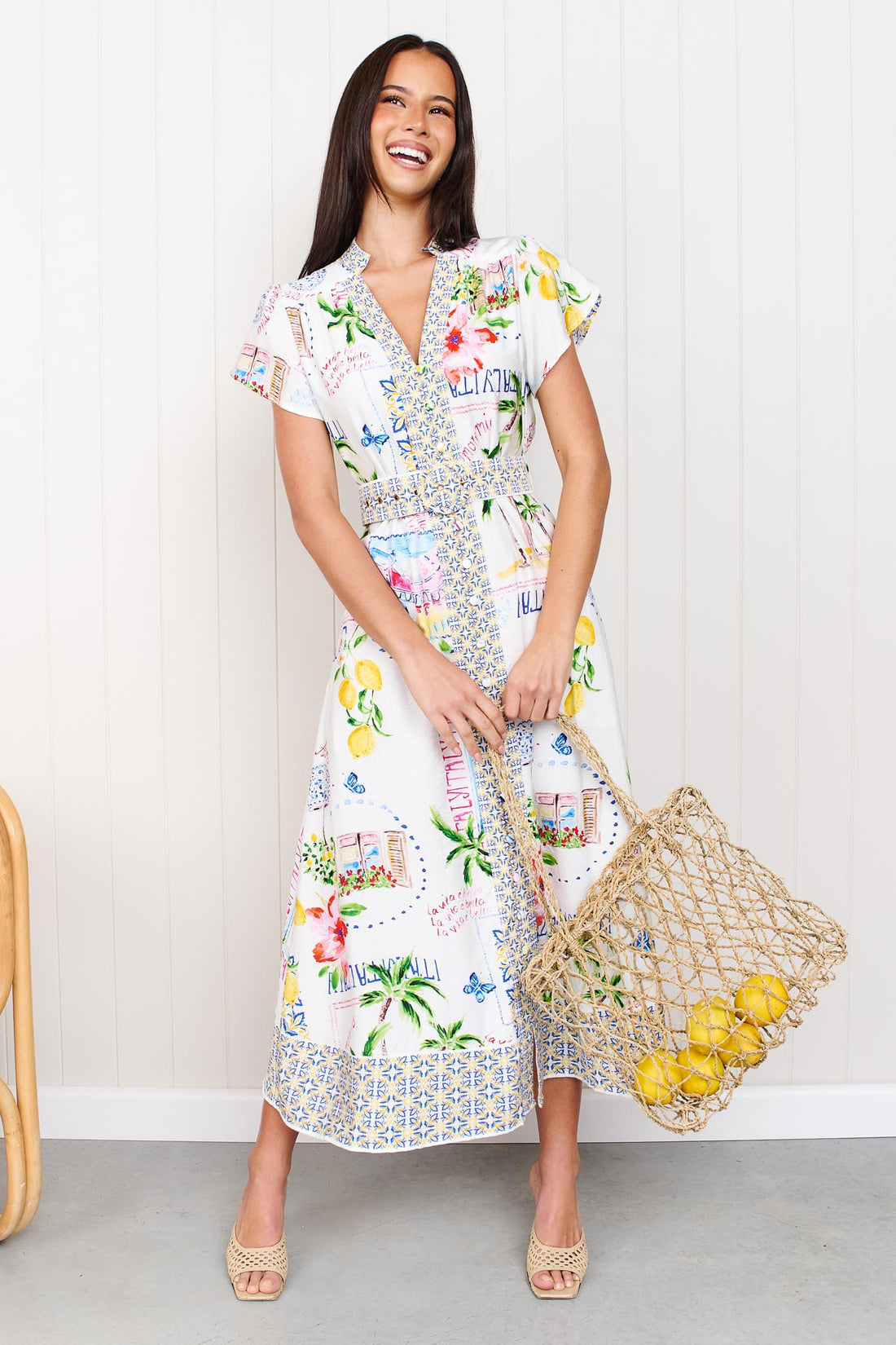 A woman wearing the minnie + MAXXIE Positano Midi Dress, featuring a colourful mediterranean-themed print, short flutter sleeves, a V-neckline, and a matching belt, set against a white panelled wall.
