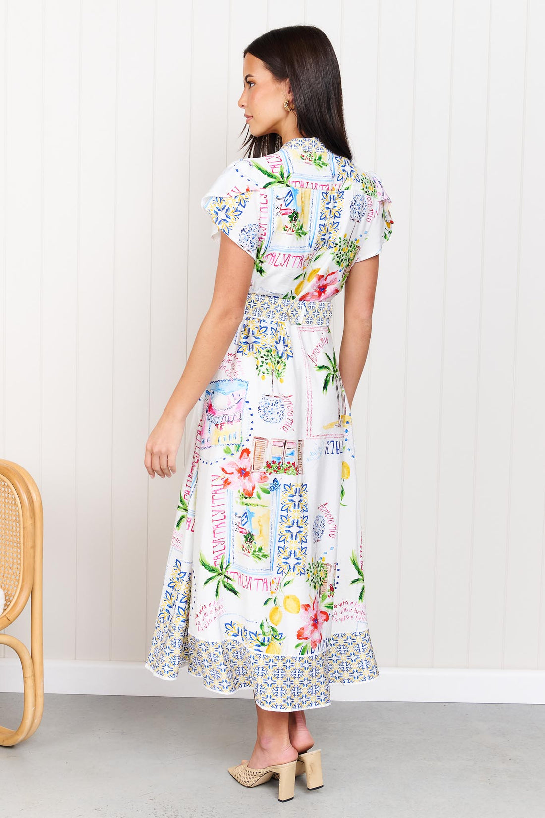 Back view of a woman wearing the minnie + MAXXIE Positano Midi Dress, featuring a colourful mediterranean-themed print, short flutter sleeves, a V-neckline, and a matching belt, set against a white panelled wall.