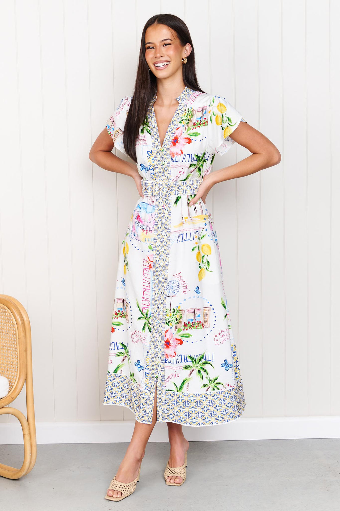 A woman wearing the minnie + MAXXIE Positano Midi Dress, featuring a colourful mediterranean-themed print, short flutter sleeves, a V-neckline, and a matching belt, set against a white panelled wall.
