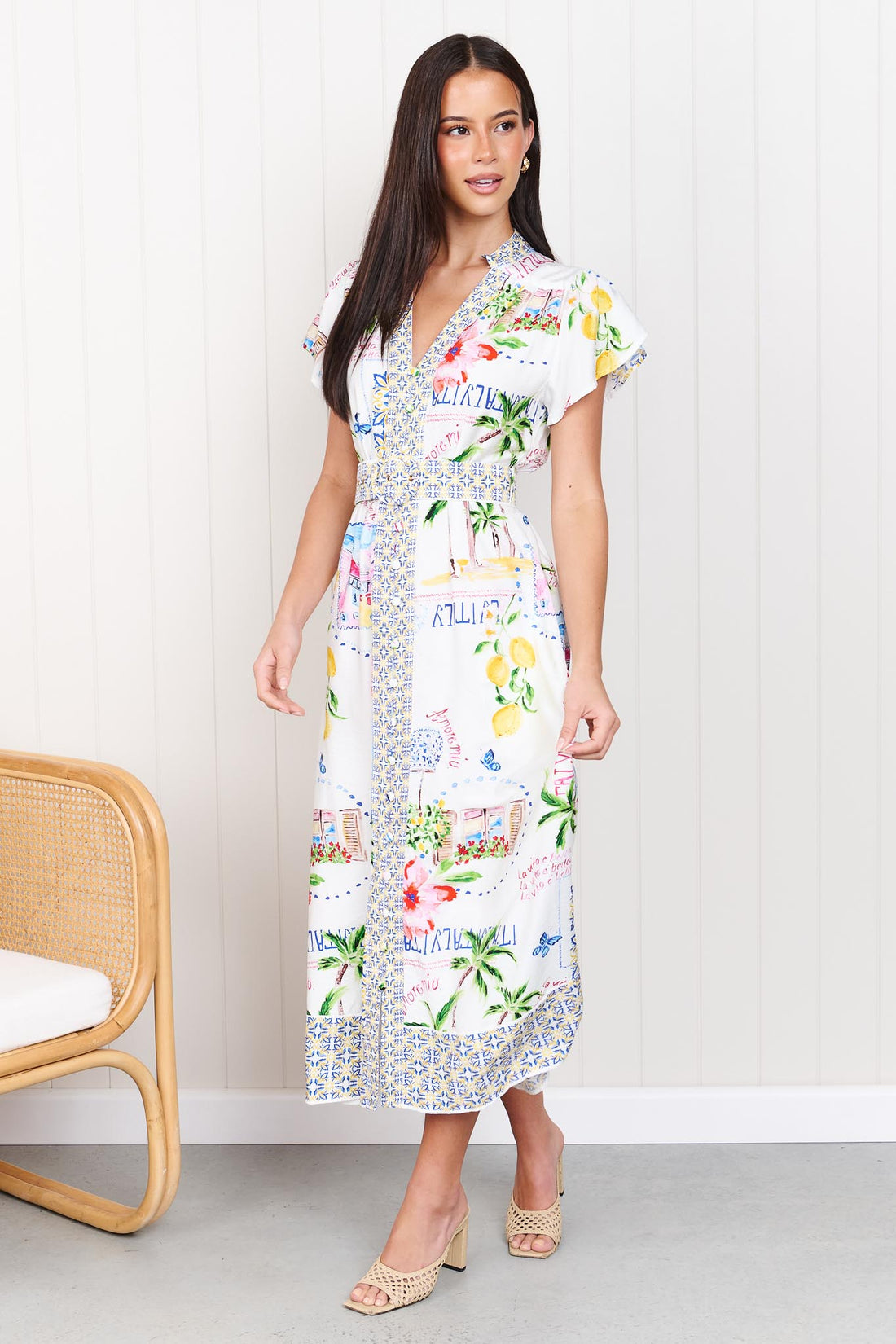 A woman wearing the minnie + MAXXIE Positano Midi Dress, featuring a colourful mediterranean-themed print, short flutter sleeves, a V-neckline, and a matching belt, set against a white panelled wall.