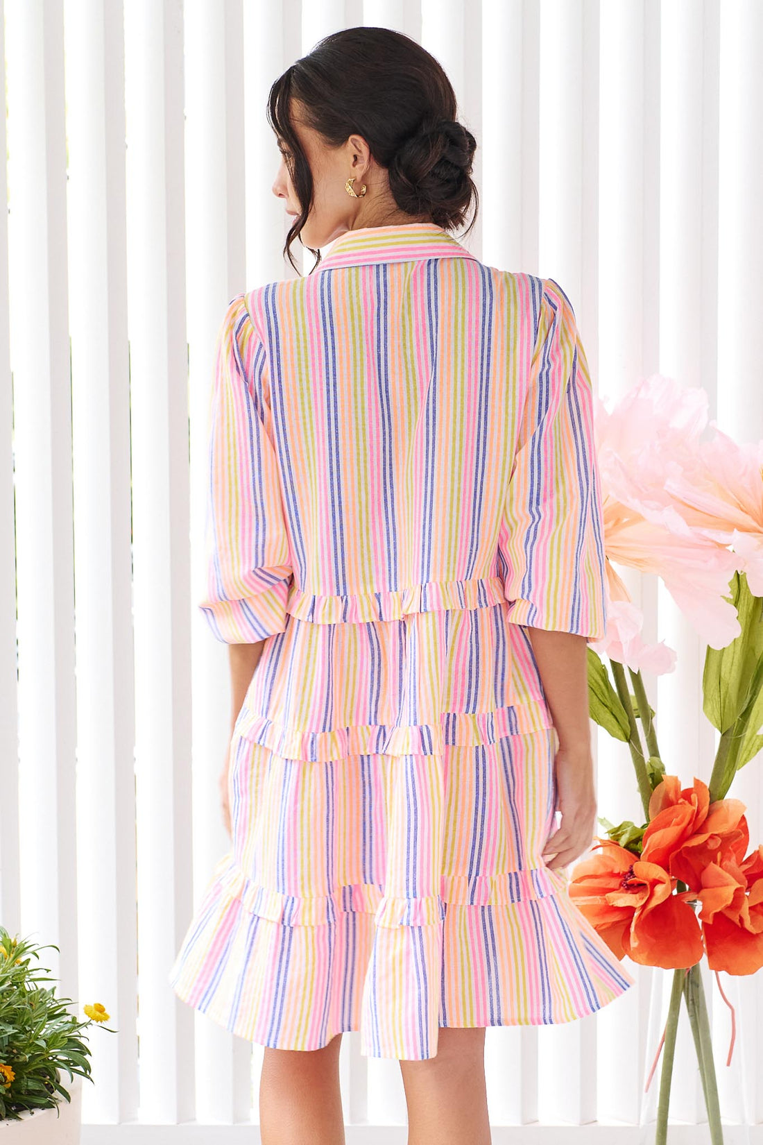 Back of a woman wearing the minnie + MAXXIE Rio A-line short dress with elbow length sleeves, a tiered shape and a colourful neon stripe print.
