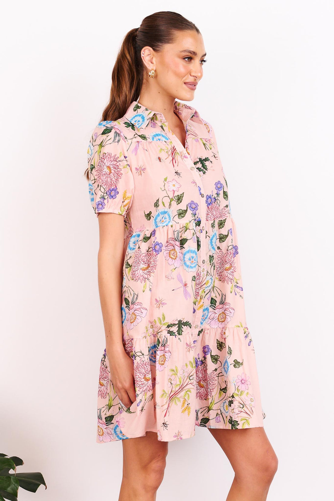 A woman wearing the minnie + MAXXIE Rosa dusty pink A-line shirt dress with short-sleeves, a collared neckline and a floral print, standing in front of a white wall.