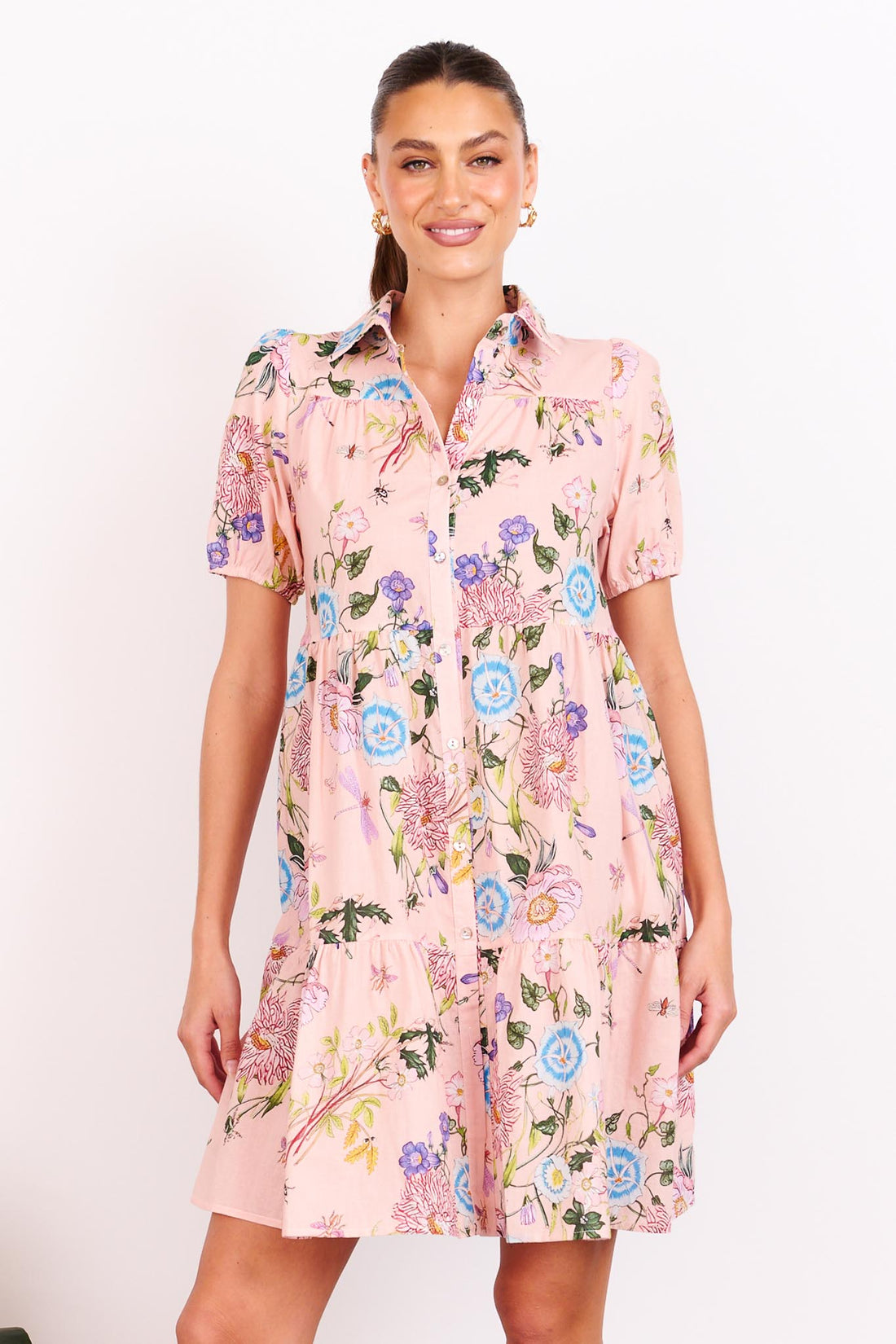 A woman wearing the minnie + MAXXIE Rosa dusty pink A-line shirt dress with short-sleeves, a collared neckline and a floral print, standing in front of a white wall.