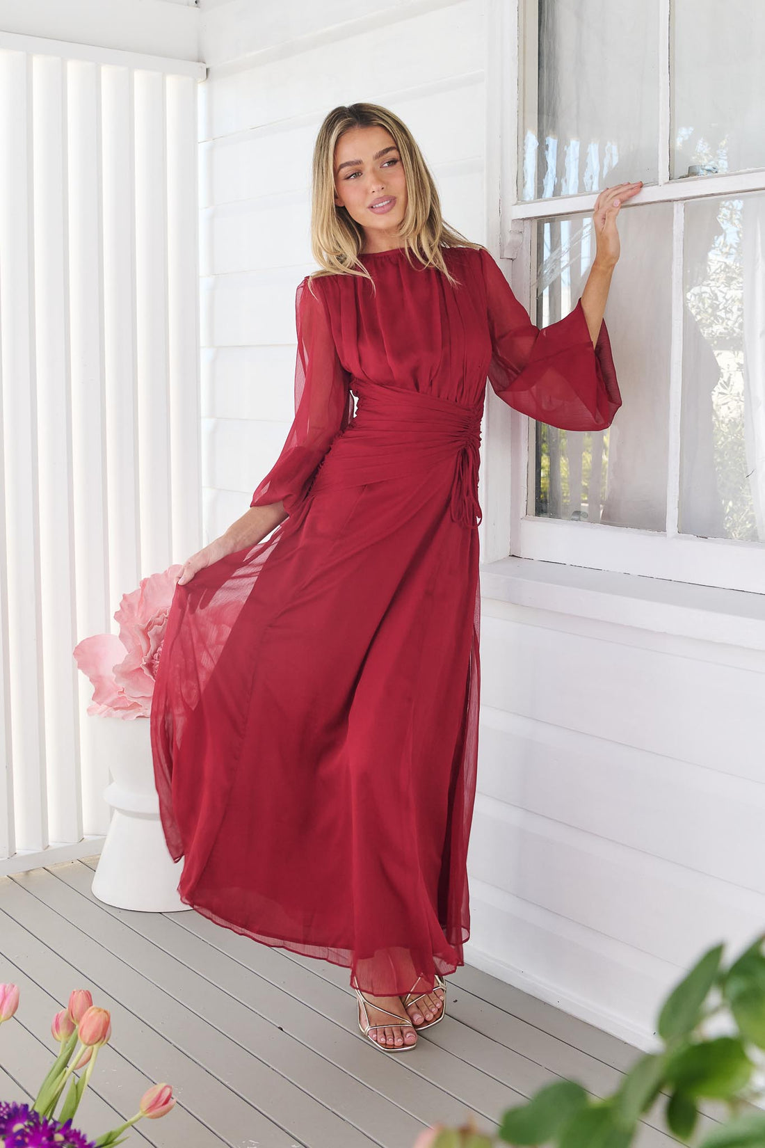 A woman wearing the minnie + MAXXIE Sabine Maxi Dress in ruby red with sheer balloon sleeves and a gathered waist tie, standing in front of a white panelled wall.
