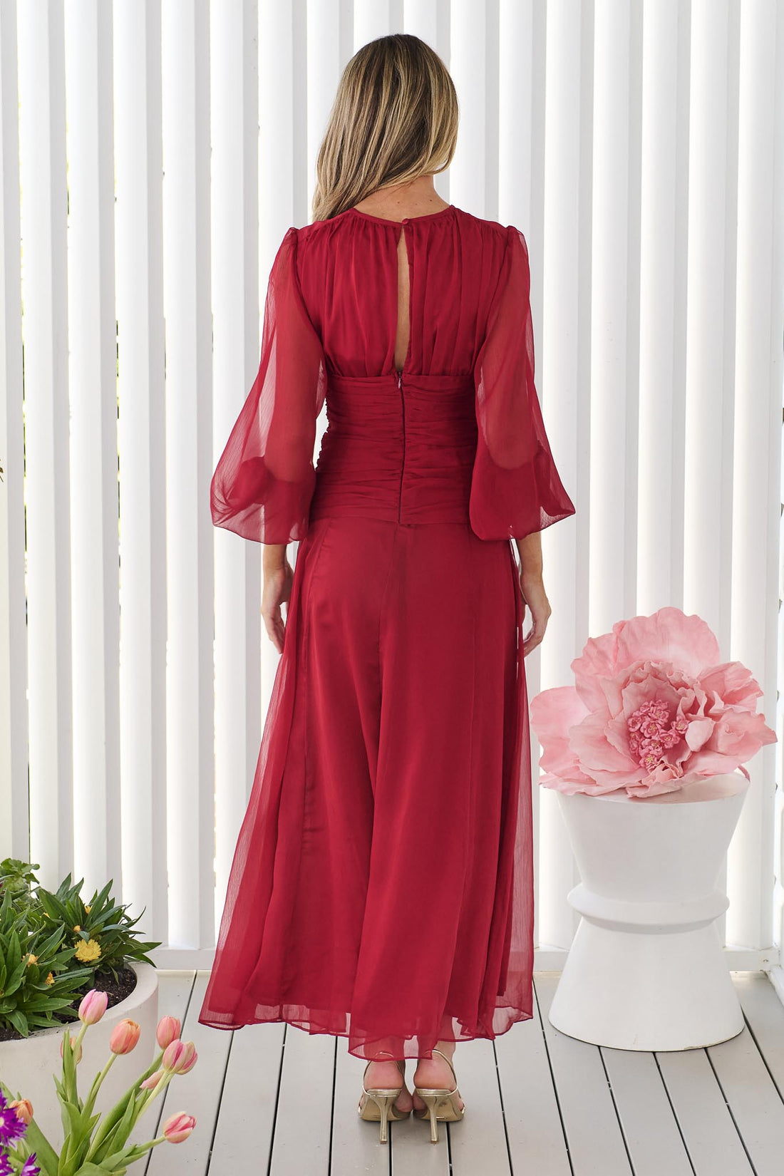 Back view of a woman wearing the minnie + MAXXIE Sabine Maxi Dress in ruby red with sheer balloon sleeves and a gathered waist tie, standing in front of a white panelled wall.