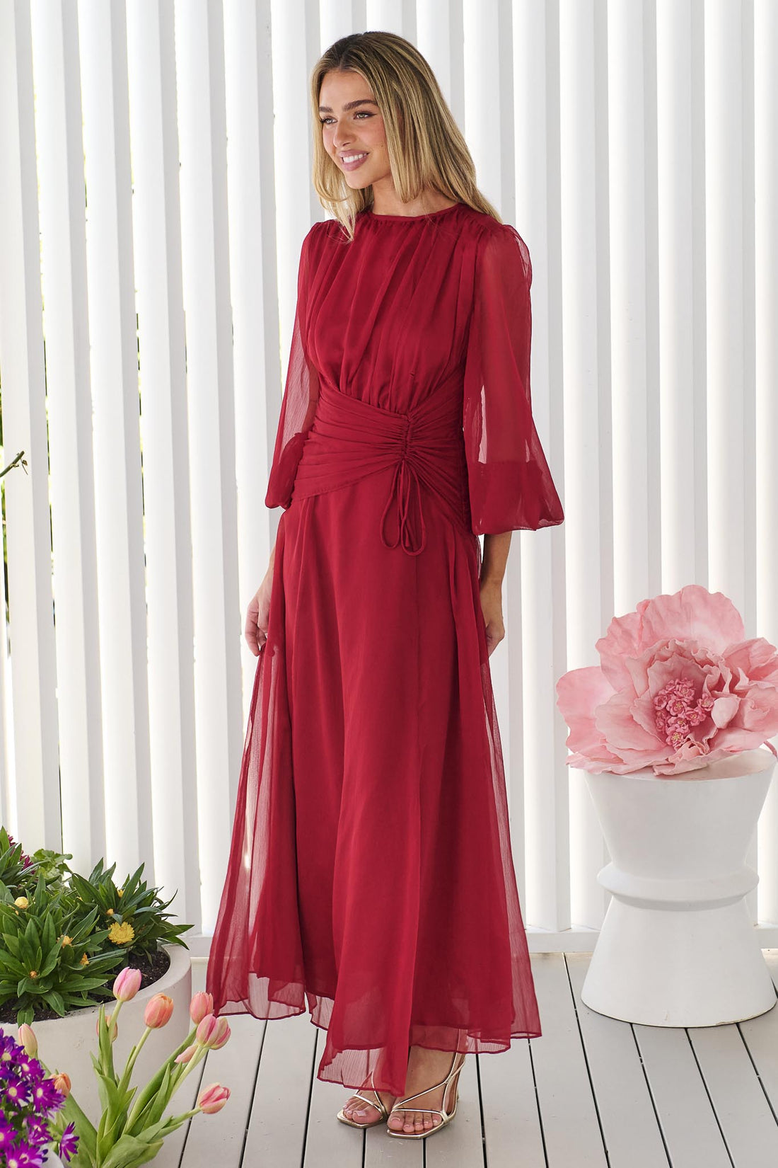 A woman wearing the minnie + MAXXIE Sabine Maxi Dress in ruby red with sheer balloon sleeves and a gathered waist tie, standing in front of a white panelled wall.
