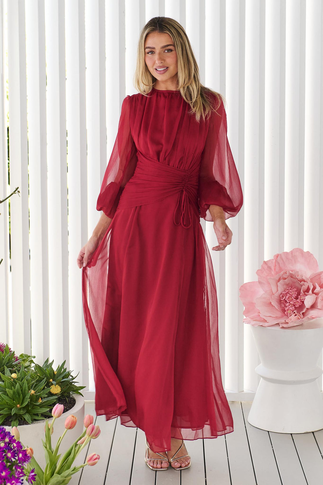 A woman wearing the minnie + MAXXIE Sabine Maxi Dress in ruby red with sheer balloon sleeves and a gathered waist tie, standing in front of a white panelled wall.