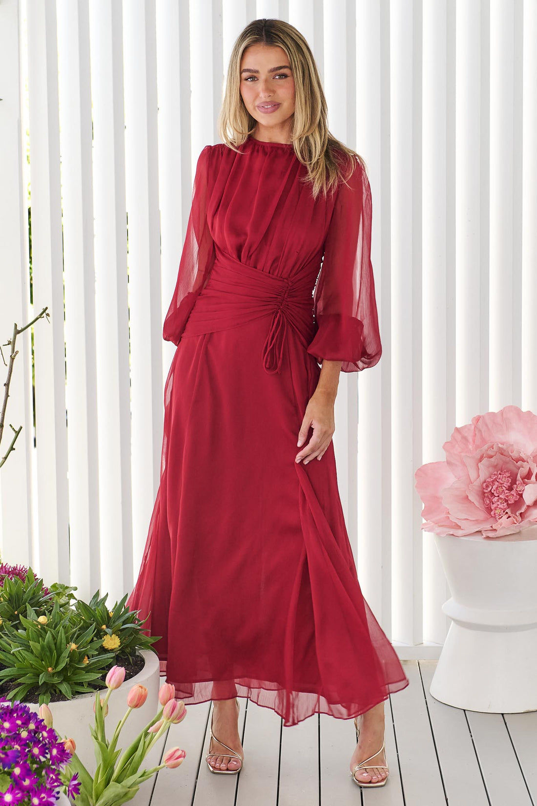 A woman wearing the minnie + MAXXIE Sabine Maxi Dress in ruby red with sheer balloon sleeves and a gathered waist tie, standing in front of a white panelled wall.