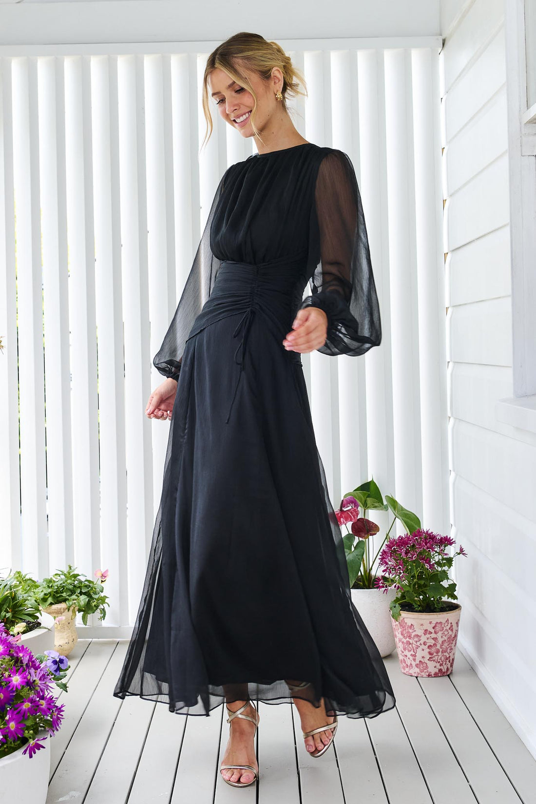 A woman wearing the minnie + MAXXIE Sabine chiffon maxi event dress in black, standing in front of a white panelled wall.