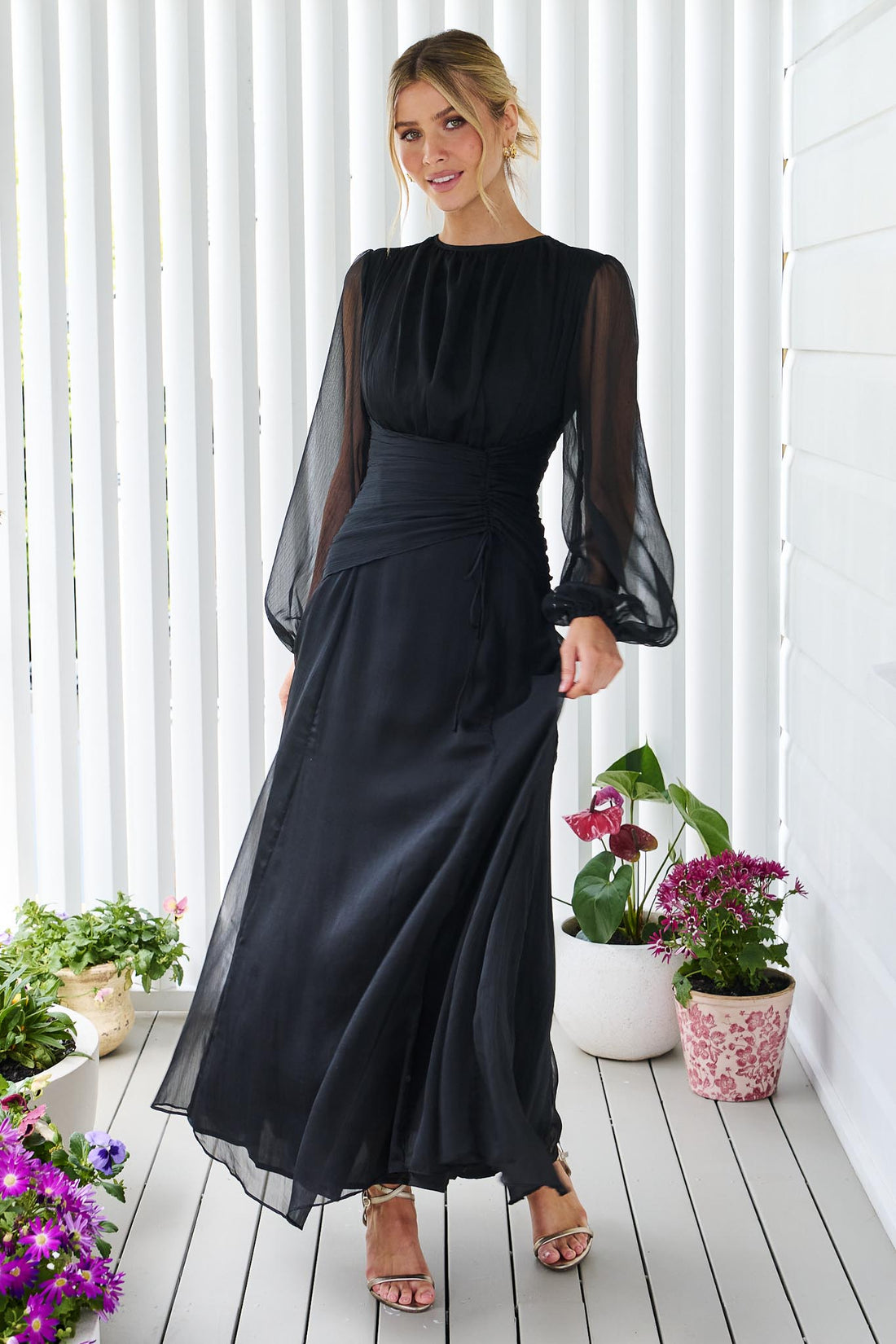 A woman wearing the minnie + MAXXIE Sabine chiffon maxi event dress in black, standing in front of a white panelled wall.