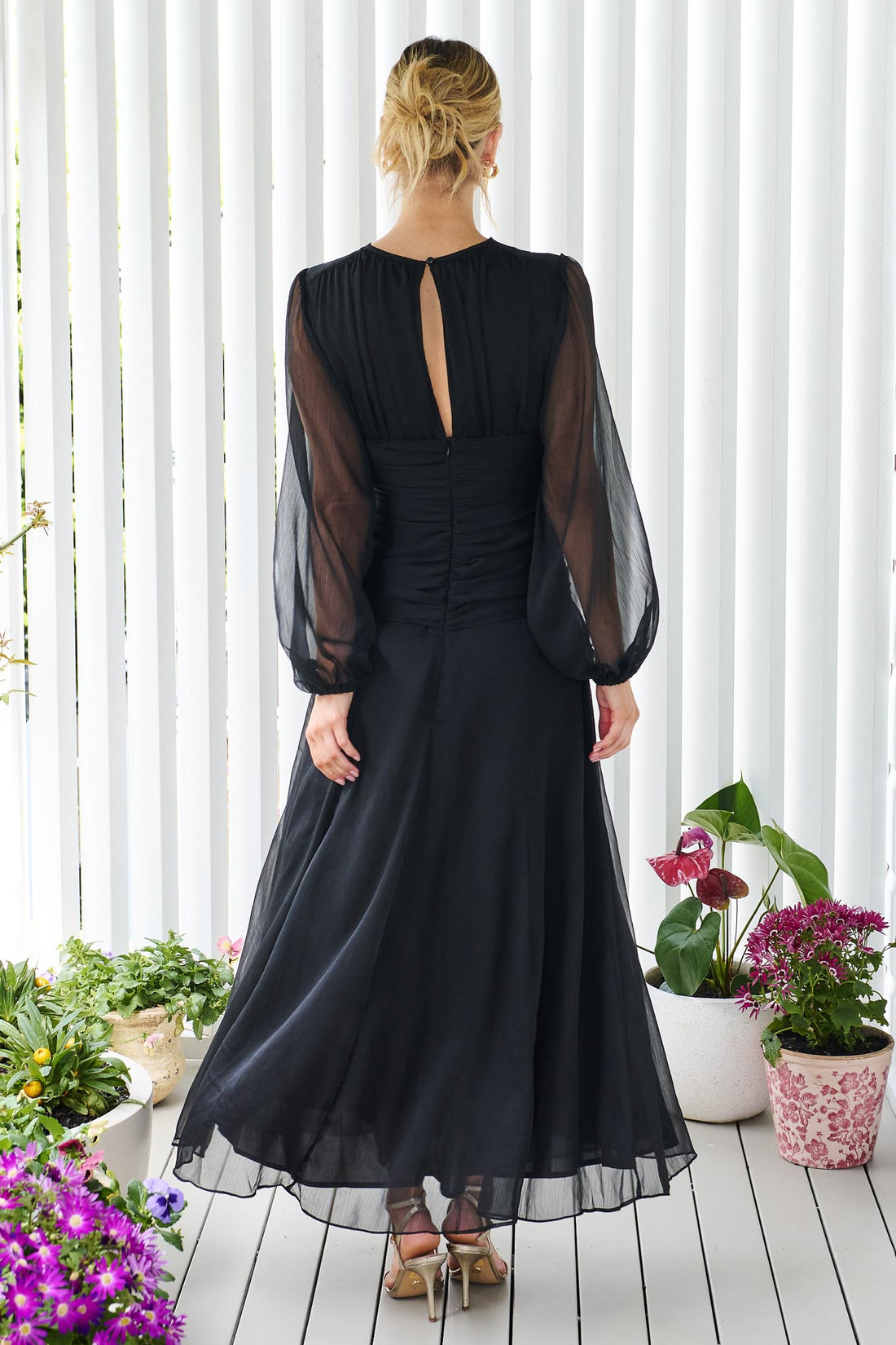 Back view of a woman wearing the minnie + MAXXIE Sabine chiffon maxi event dress in black, standing in front of a white panelled wall.