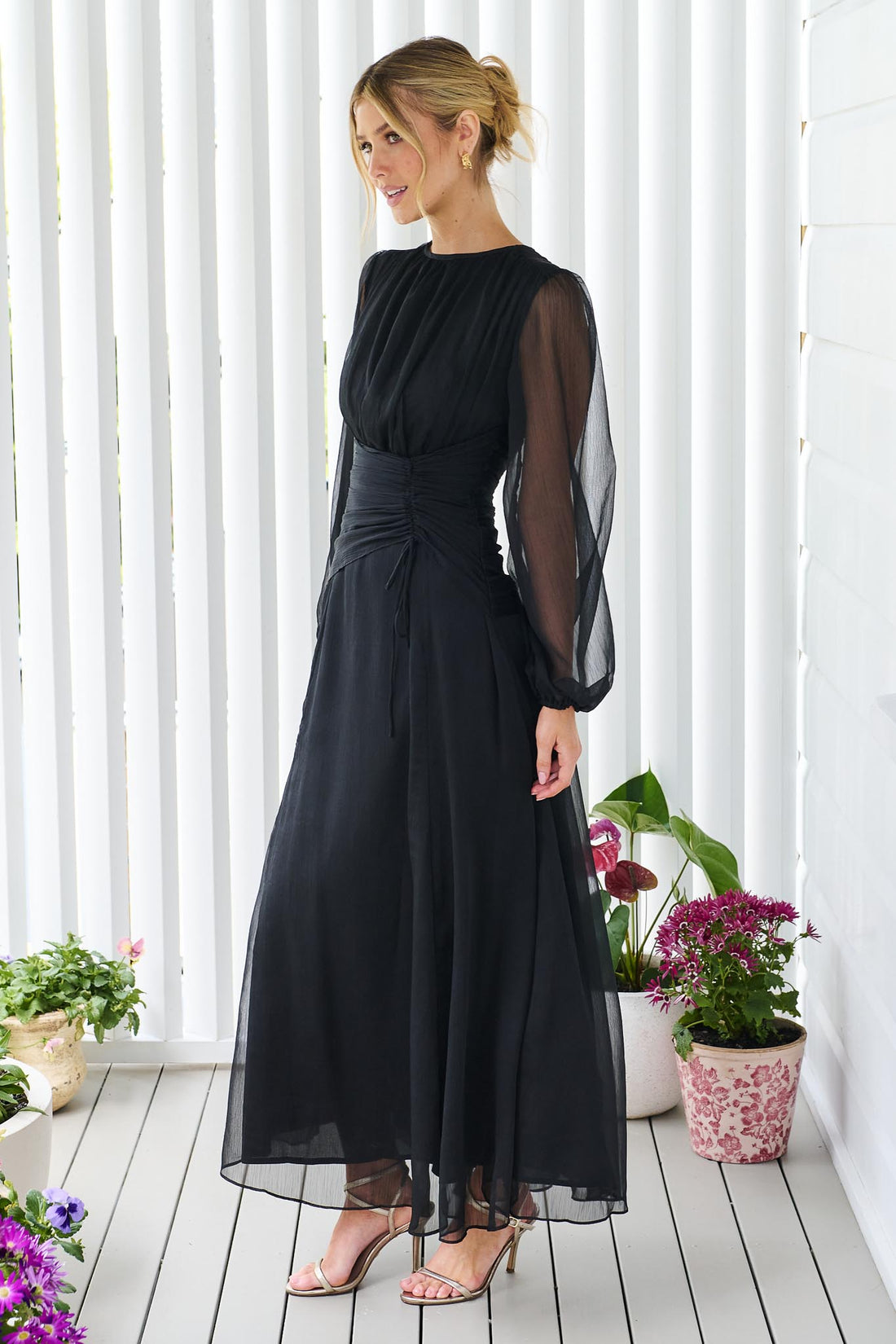 Side view of a woman wearing the minnie + MAXXIE Sabine chiffon maxi event dress in black, standing in front of a white panelled wall.