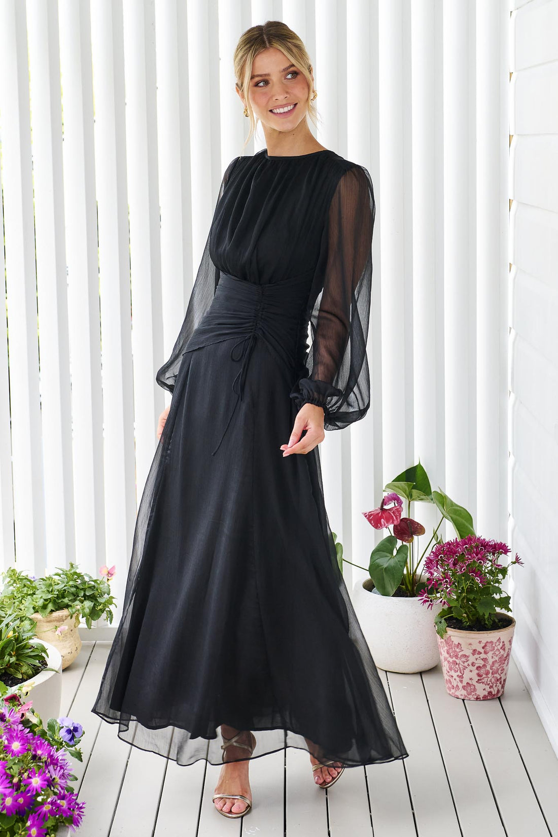 Side view of a woman wearing the minnie + MAXXIE Sabine chiffon maxi event dress in black, standing in front of a white panelled wall.