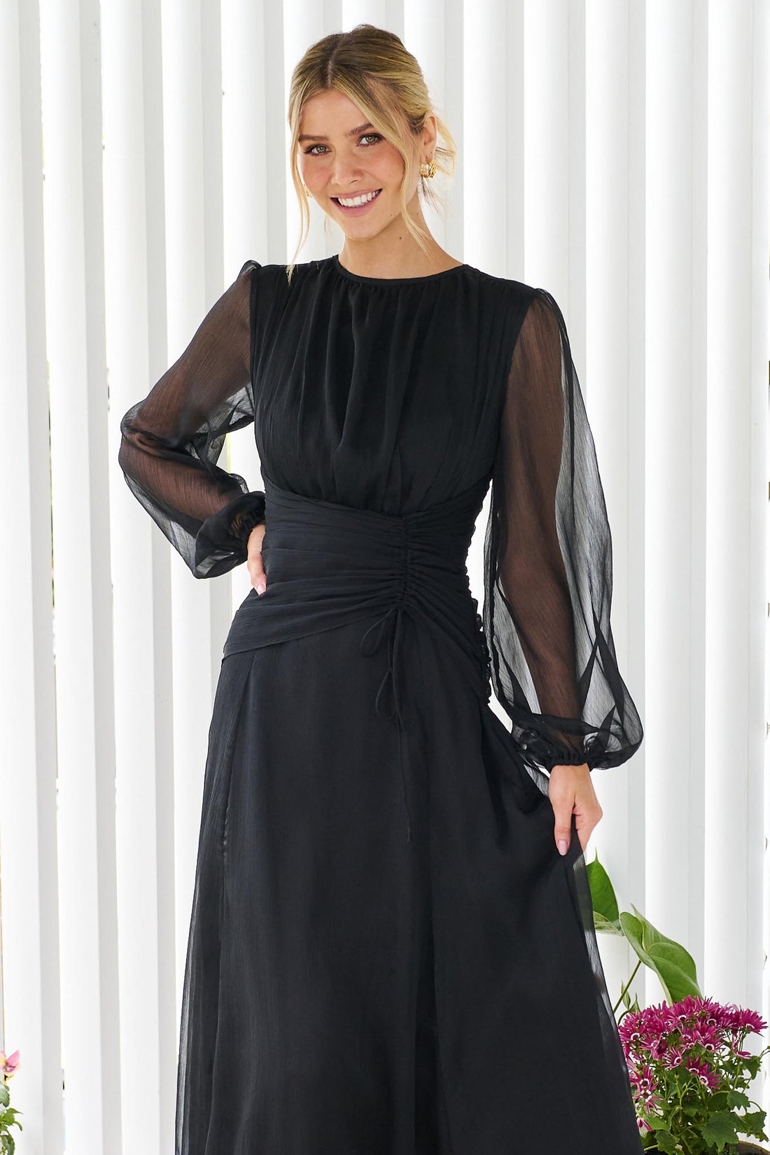 Close up of a woman wearing the minnie + MAXXIE Sabine chiffon maxi event dress in black, standing in front of a white panelled wall.