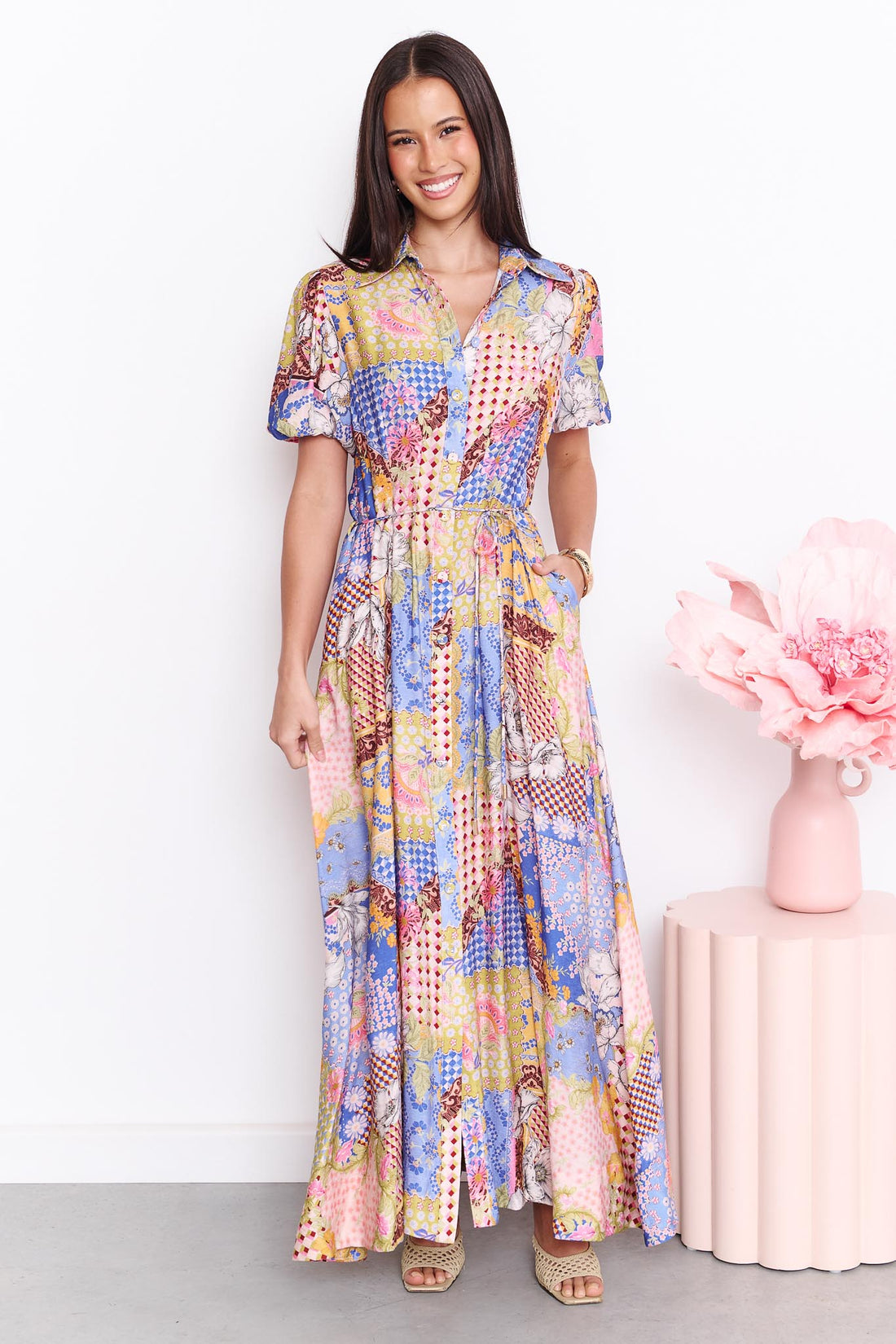 A woman wearing the minnie + MAXXIE Sabrina short-sleeved maxi dress with a colourful patchwork print. The dress features a mix of blue, pink, yellow, and white patterns.