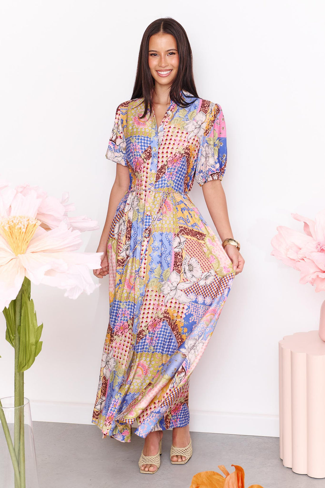 A woman wearing the minnie + MAXXIE Sabrina short-sleeved maxi dress with a colourful patchwork print, surrounded by large paper flowers. The dress features a mix of blue, pink, yellow, and white patterns.