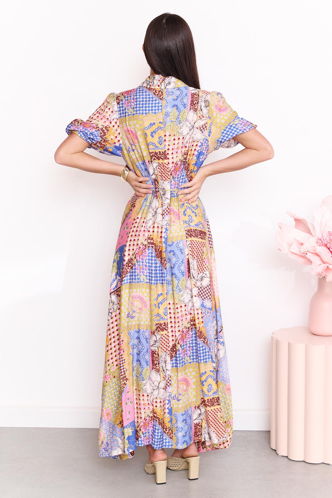 Back view of a woman wearing the minnie + MAXXIE Sabrina short-sleeved maxi dress with a colourful patchwork print. The dress features a mix of blue, pink, yellow, and white patterns.