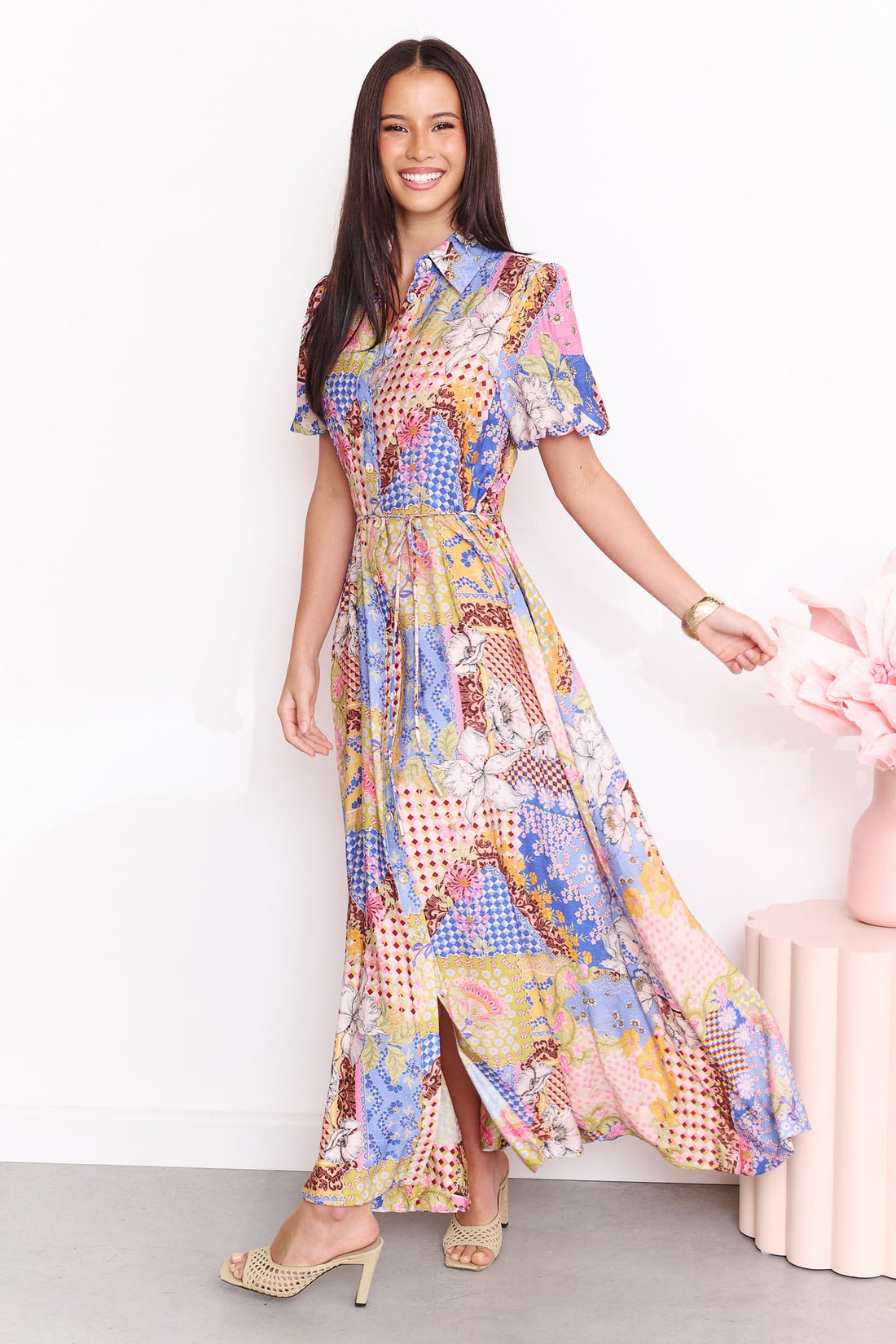 A woman wearing the minnie + MAXXIE Sabrina short-sleeved maxi dress with a colourful patchwork print. The dress features a mix of blue, pink, yellow, and white patterns.