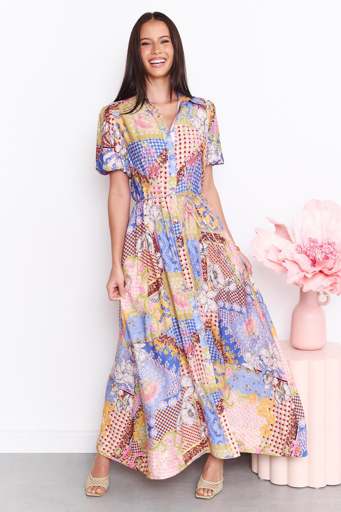 A woman wearing the minnie + MAXXIE Sabrina short-sleeved maxi dress with a colourful patchwork print. The dress features a mix of blue, pink, yellow, and white patterns.
