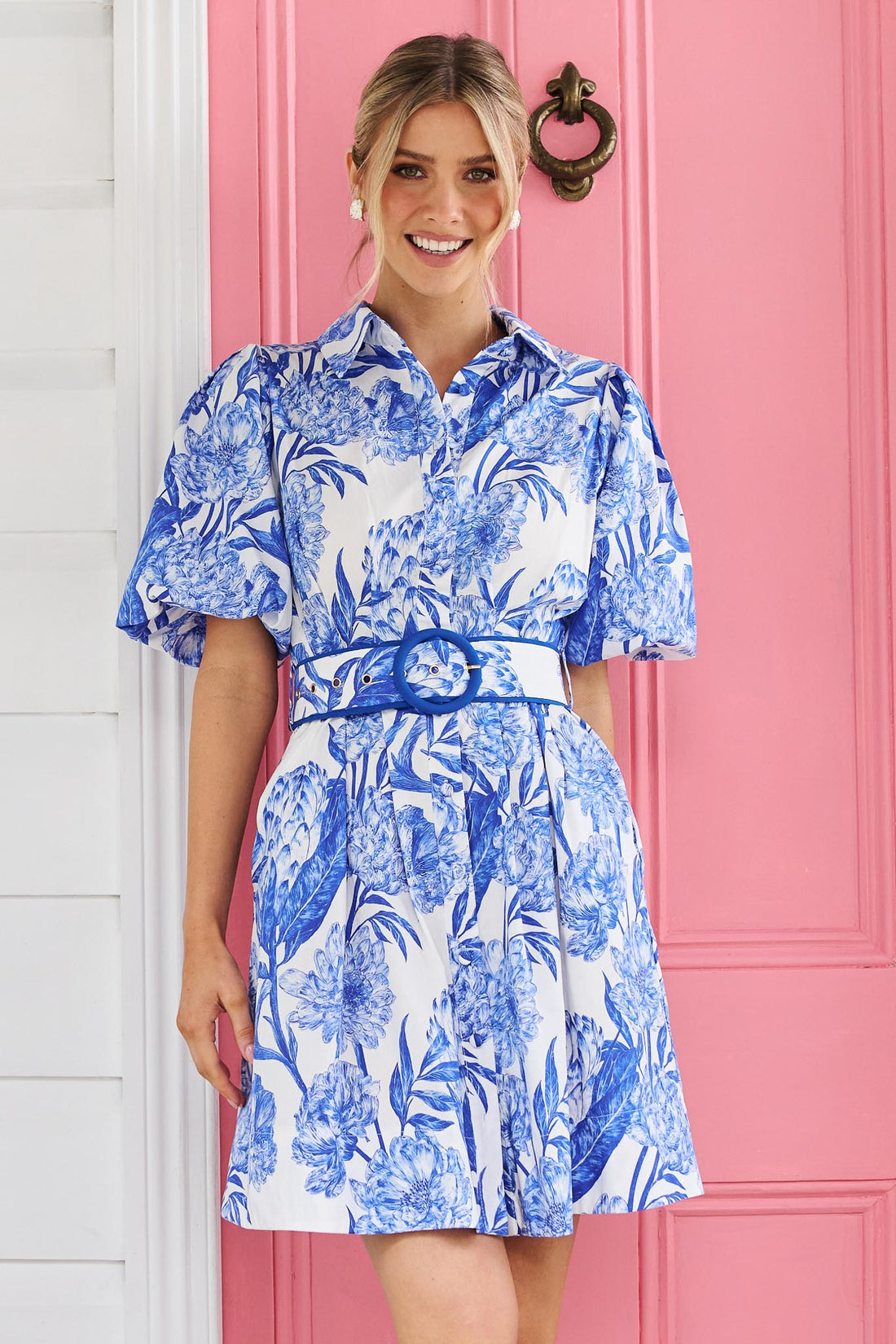 A woman wearing the minnie + MAXXIE blue and white floral print Sarita mini dress with puff sleeves and a matching belt, standing in front of a pink door.