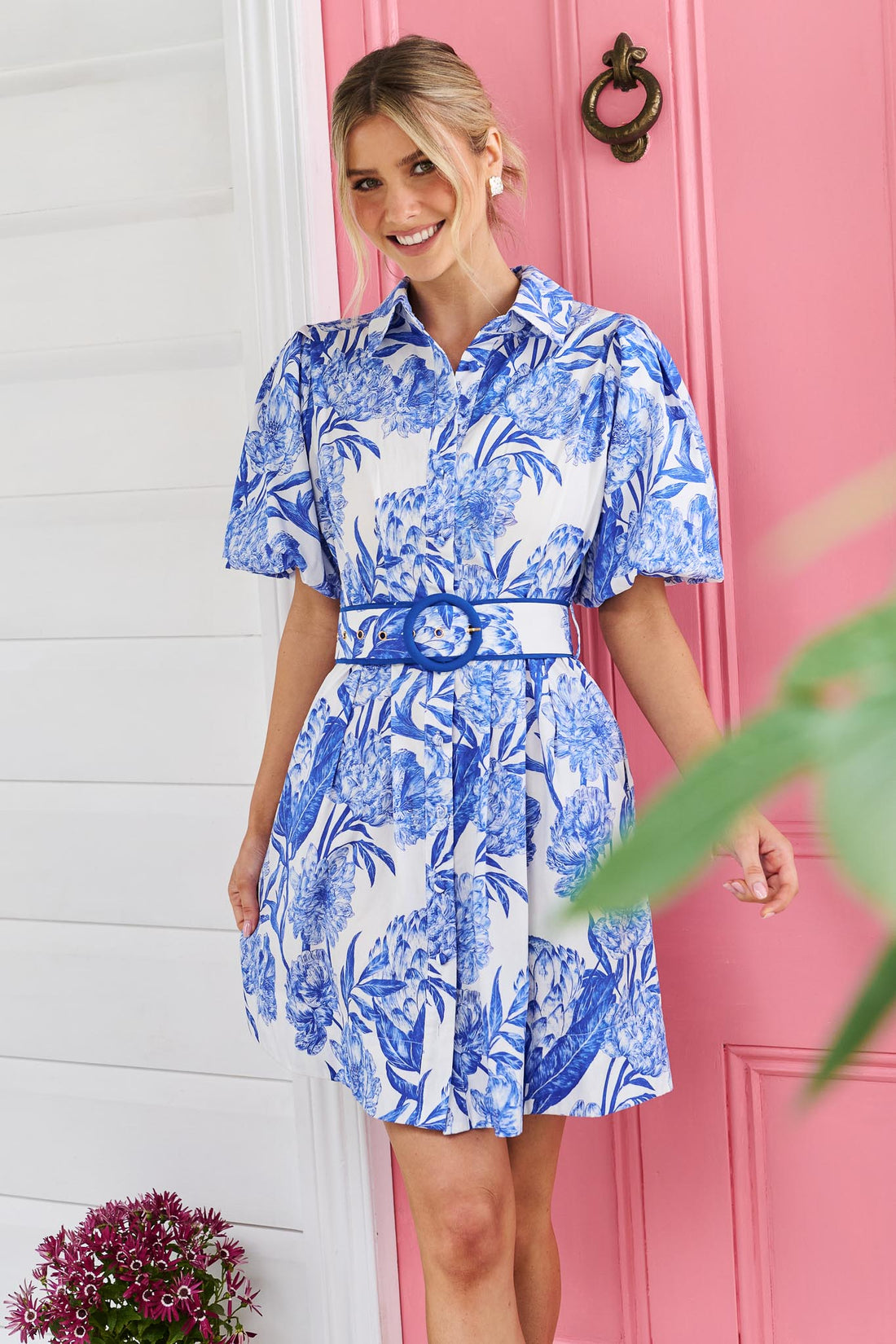 A woman wearing the minnie + MAXXIE blue and white floral print Sarita mini dress with puff sleeves and a matching belt, standing in front of a pink door.