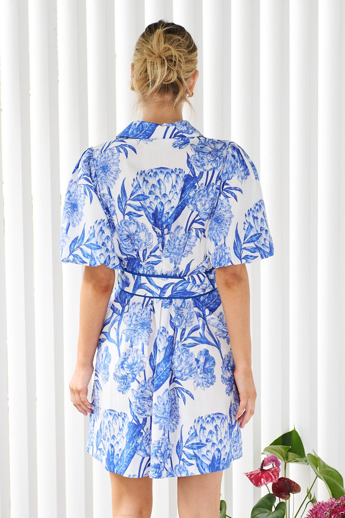 Back view of a woman wearing the minnie + MAXXIE blue and white floral print Sarita mini dress with puff sleeves and a matching belt, standing in front of a pink door.