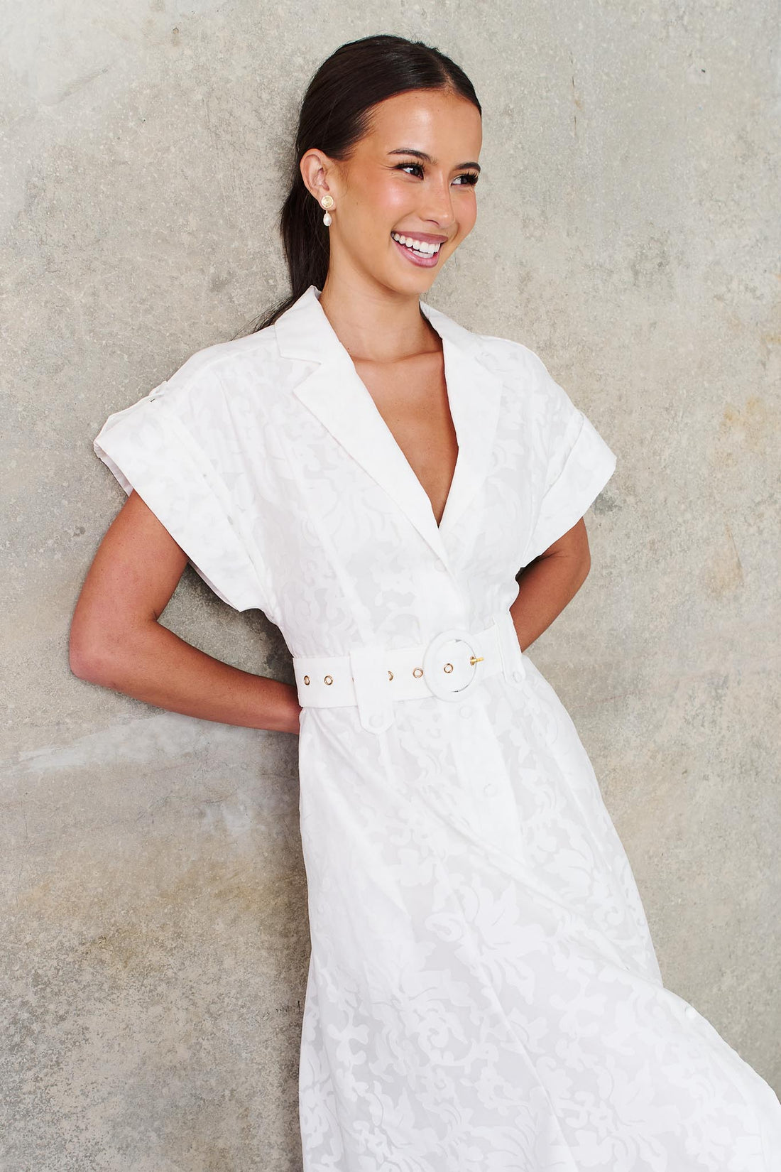 A woman wearing the minnie + MAXXIE Savannah white midi dress with short cap sleeves, a deep V-neckline and a matching belt, standing against a textured concrete wall.