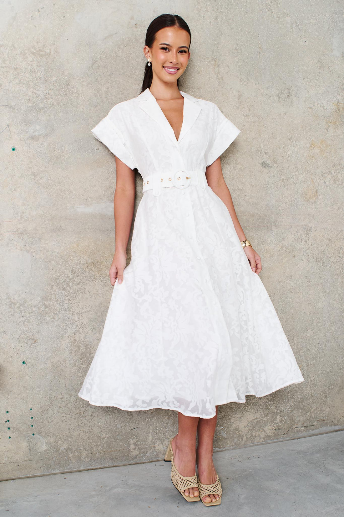 A woman wearing the minnie + MAXXIE Savannah white midi dress with short cap sleeves, a deep V-neckline and a matching belt, standing against a textured concrete wall.
