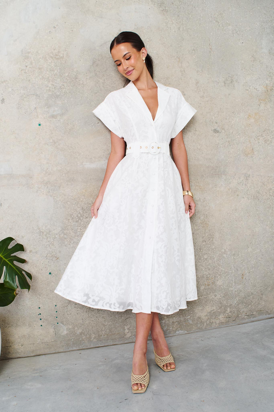 A woman wearing the minnie + MAXXIE Savannah white midi dress with short cap sleeves, a deep V-neckline and a matching belt, standing against a textured concrete wall.