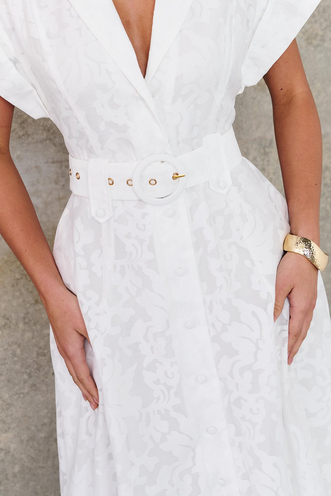 Close-up of a woman wearing the minnie + MAXXIE Savannah white midi dress with short cap sleeves, a deep V-neckline and a matching belt, standing against a textured concrete wall.