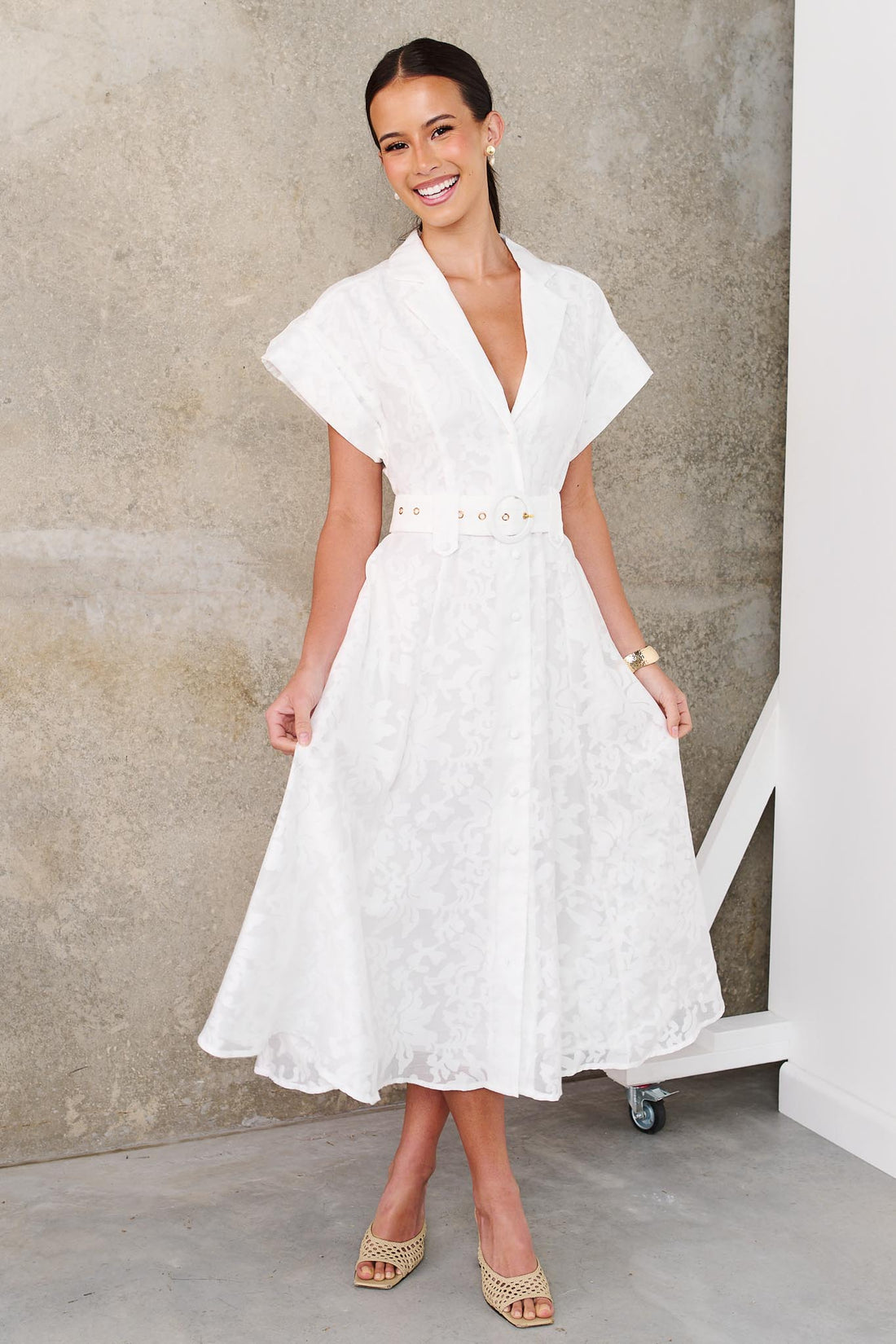 A woman wearing the minnie + MAXXIE Savannah white midi dress with short cap sleeves, a deep V-neckline and a matching belt, standing against a textured concrete wall.