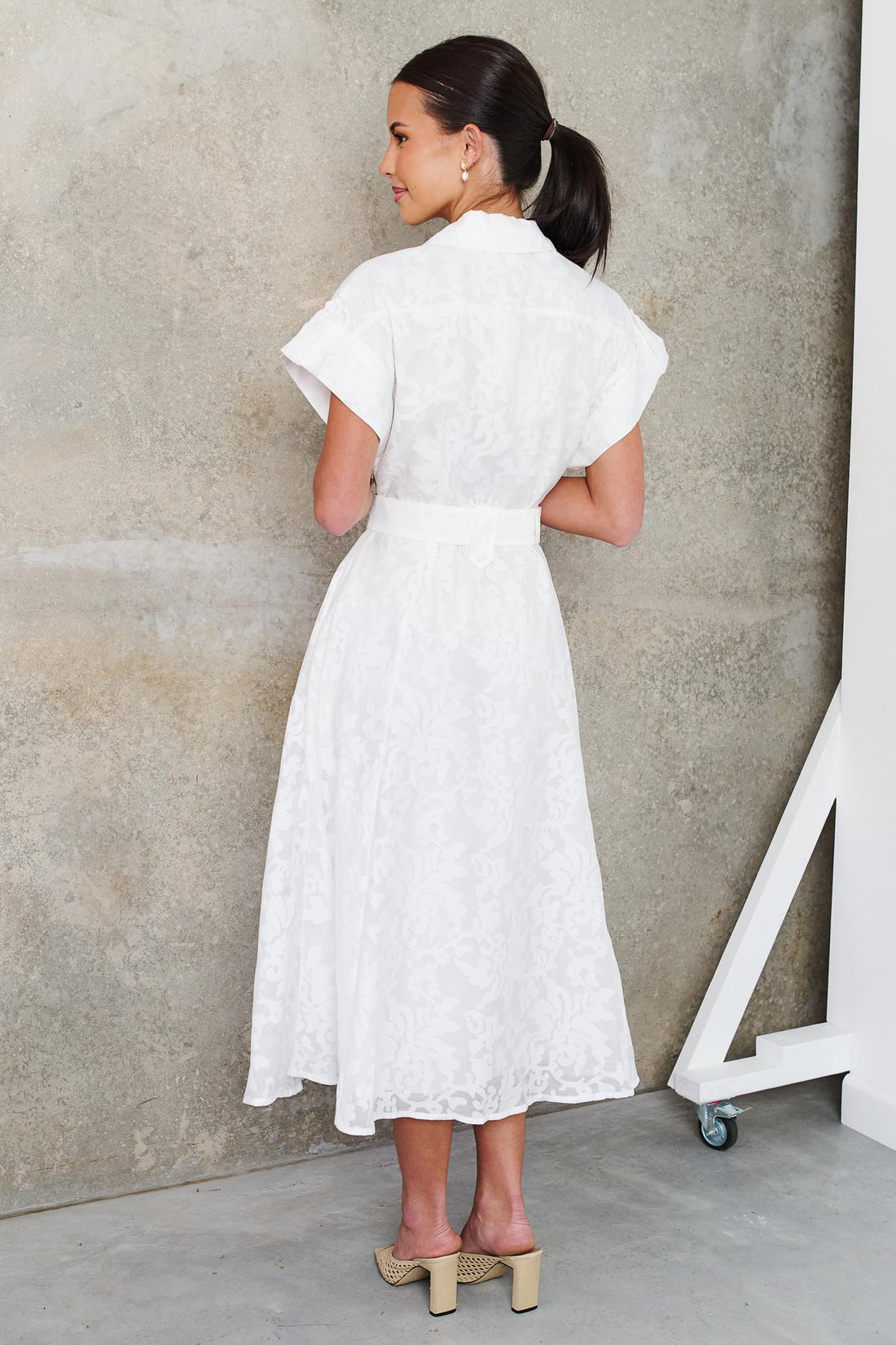 Back view of a woman wearing the minnie + MAXXIE Savannah white midi dress with short cap sleeves, a deep V-neckline and a matching belt, standing against a textured concrete wall.