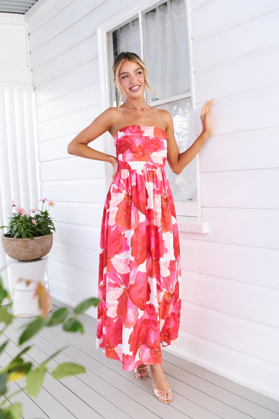 Back view of a woman wearing the minnie + MAXXIE Sienna Strapless pink floral event dress, standing against a white panelled wall with potted flowers in the background.